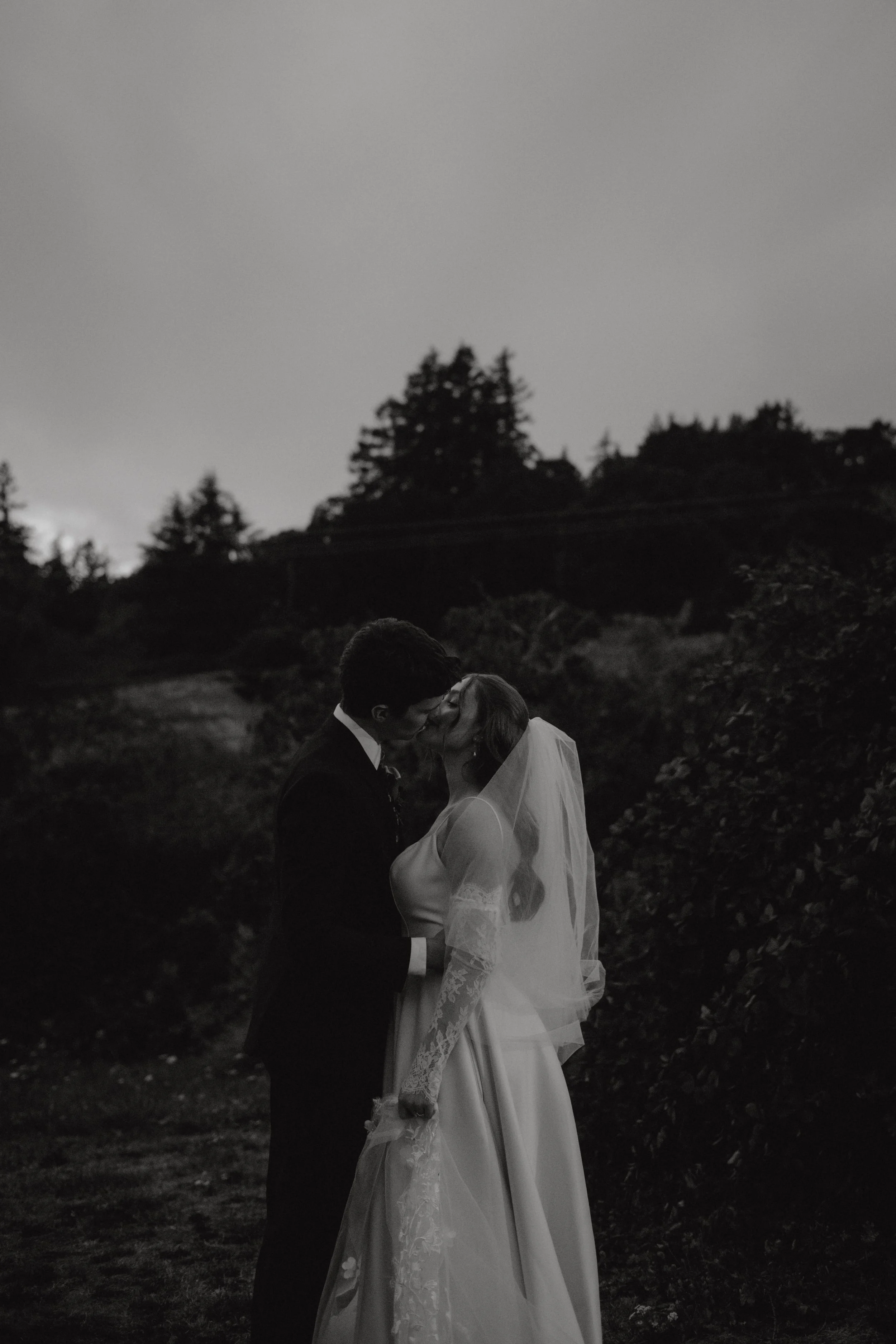 Black and white photo of a couple kissing outdoors at dusk, surrounded by trees and with a cloudy sky.