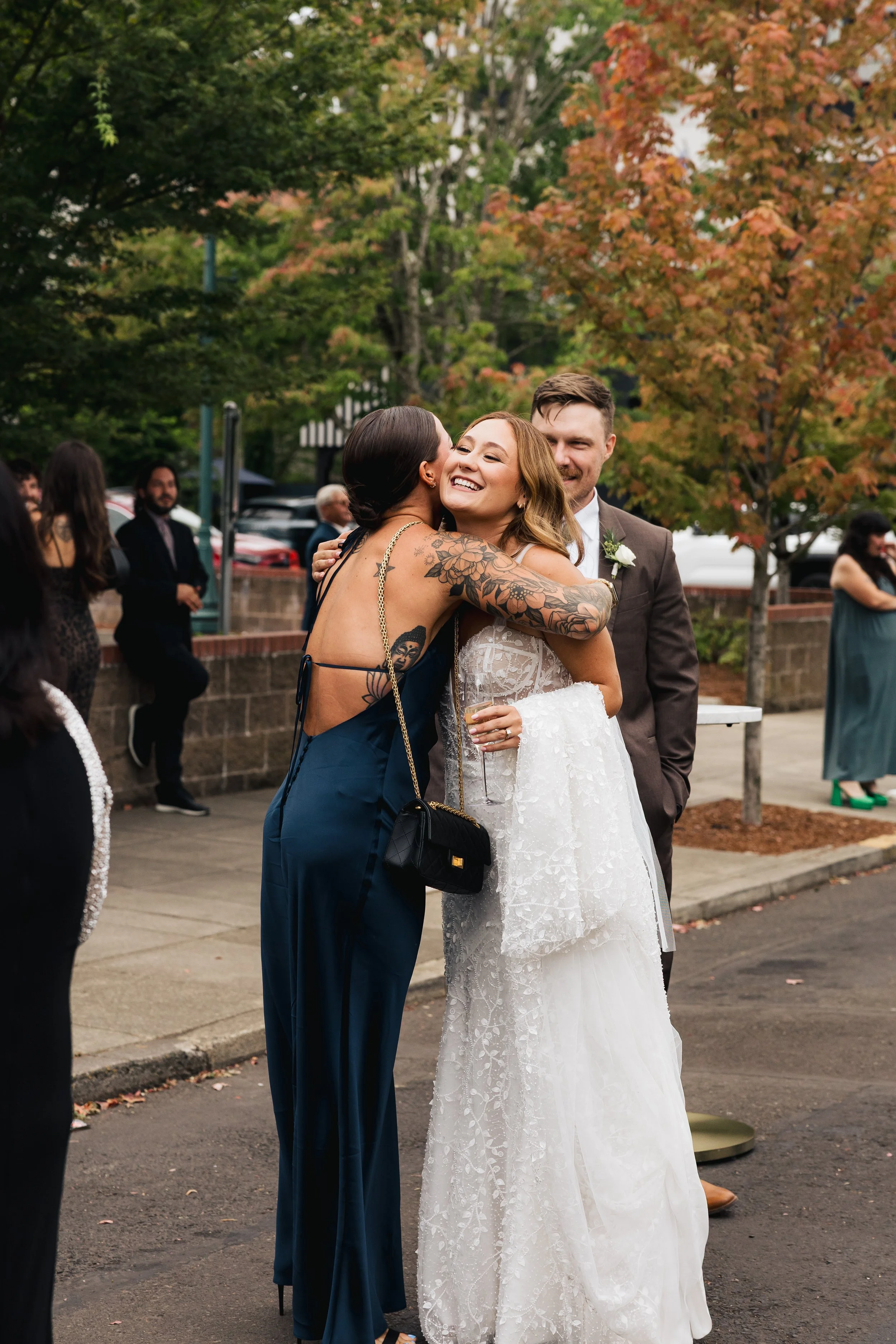 A group of people at an outdoor wedding reception during fall, with two women hugging and smiling, one wearing a white wedding dress and the other in a navy blue dress, and a man dressed in a suit nearby.