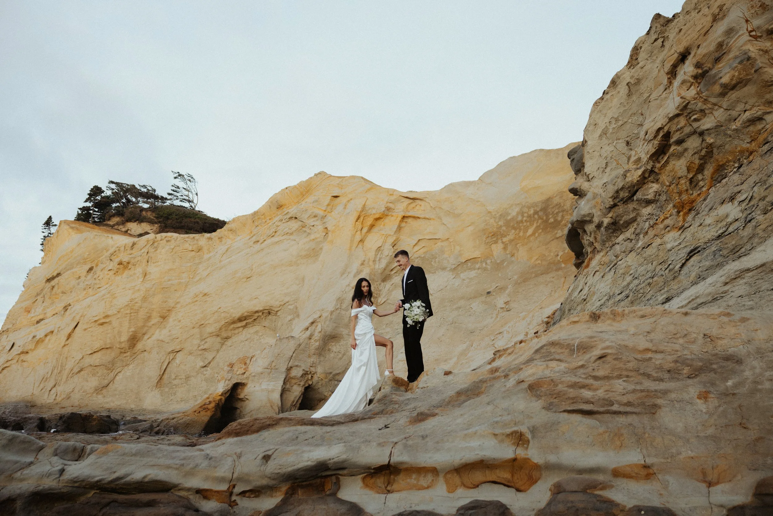 A bride and groom standing on rocks near the beach, with large yellowish cliffs behind them. The bride is wearing a white wedding dress and holding a bouquet, while the groom is in a black suit, holding her hand.