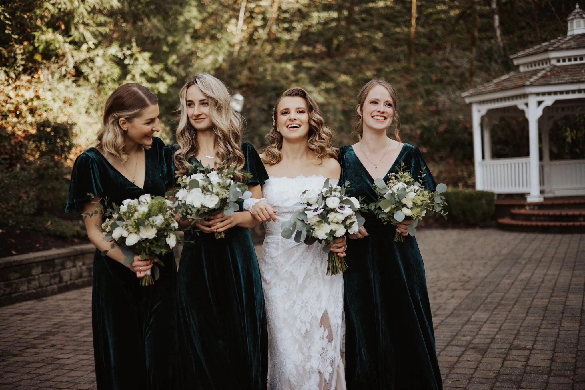 A bride in a white wedding dress walking arm-in-arm with four bridesmaids in dark green velvet dresses, all holding bouquets of white and green flowers, outdoors on a paved path with trees and a white gazebo in the background.