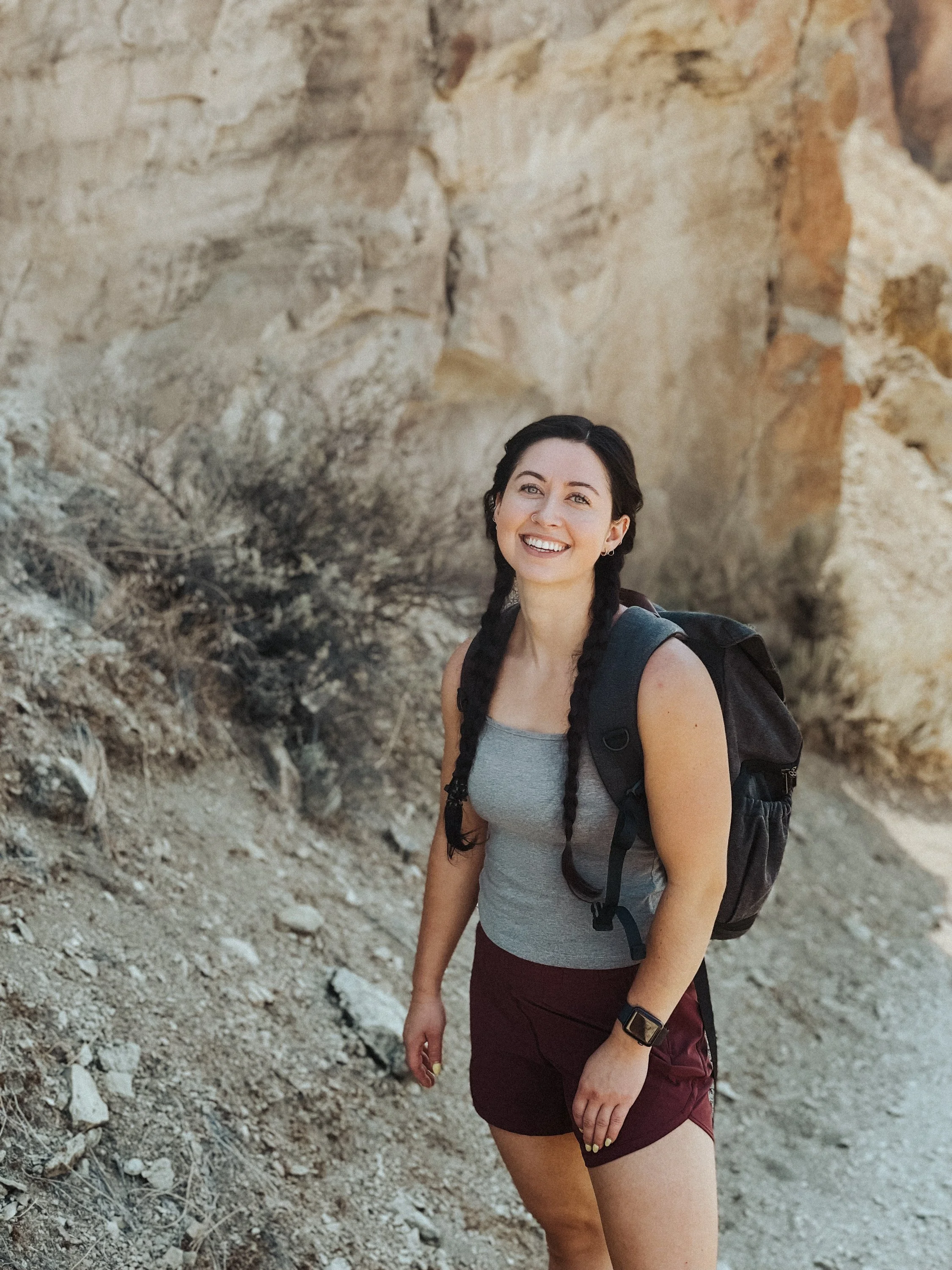 A woman with braided dark hair smiling outdoors near rocky terrain, wearing a gray sleeveless top, maroon shorts, a black backpack, and a smartwatch.