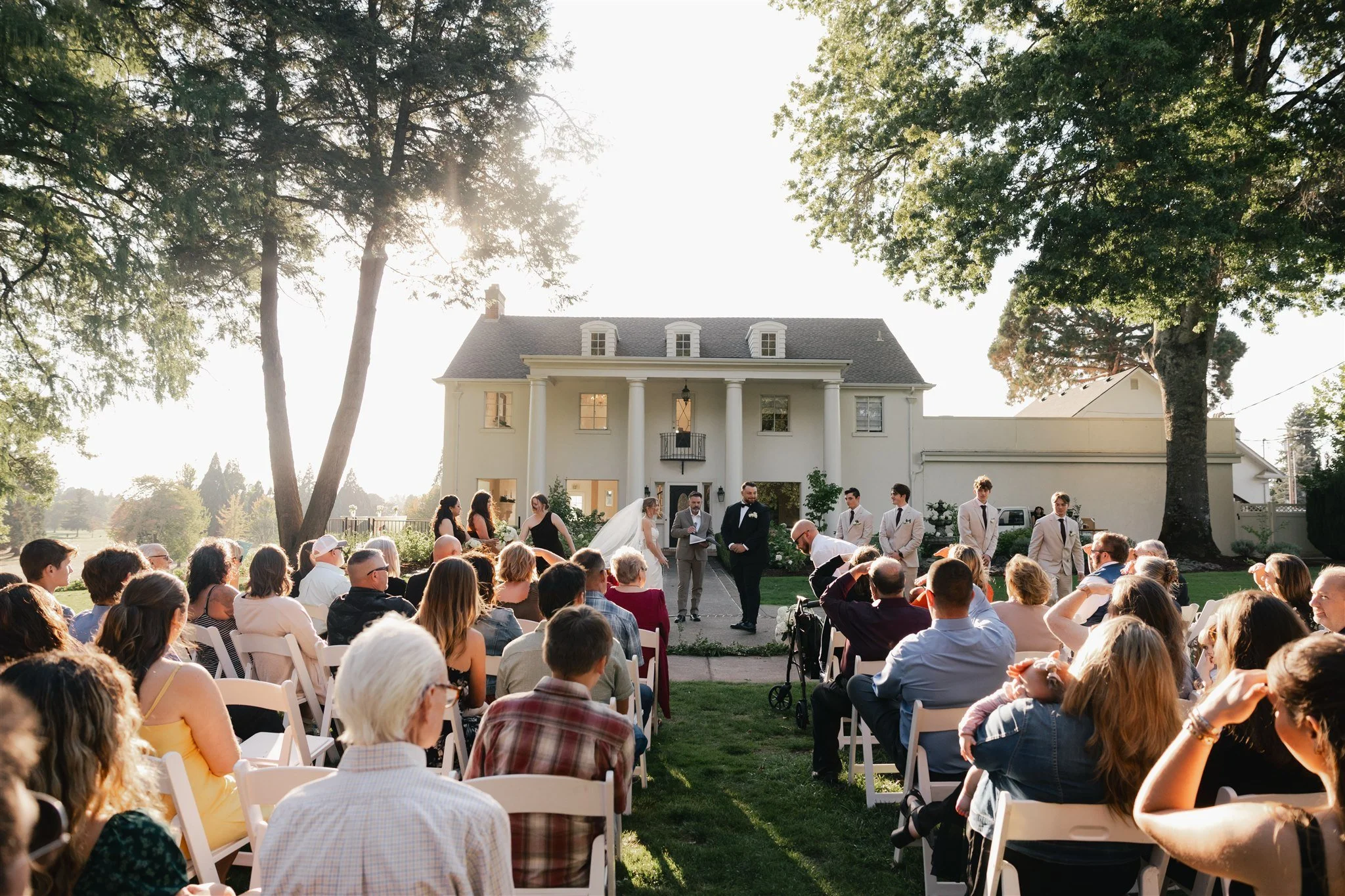 An outdoor wedding ceremony with a large crowd seated on white chairs in front of a two-story house with columns. The bride and groom are standing in front of the house, exchanging vows, with the officiant nearby. The scene is attended by guests, and