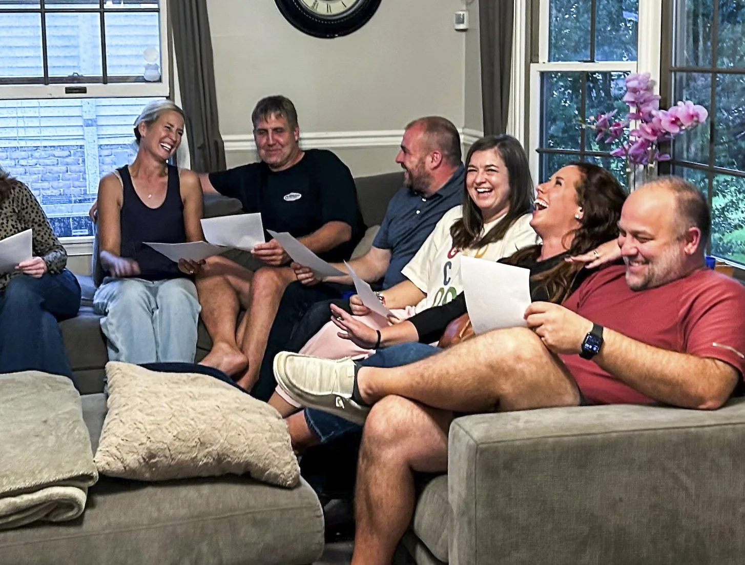 A group of six adults sitting on a couch in a living room, laughing and holding papers, engaging in a joyful activity together.