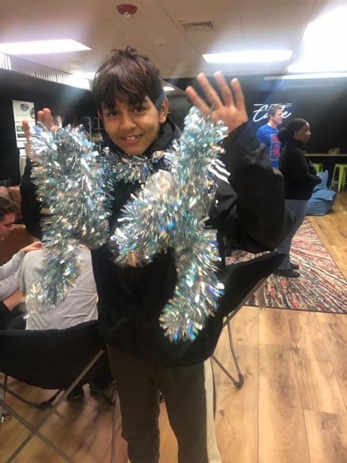 A young boy smiling and waving at the camera, wearing shiny silver tinsel around his neck and shoulders, in an indoor setting with other people sitting and standing in the background.