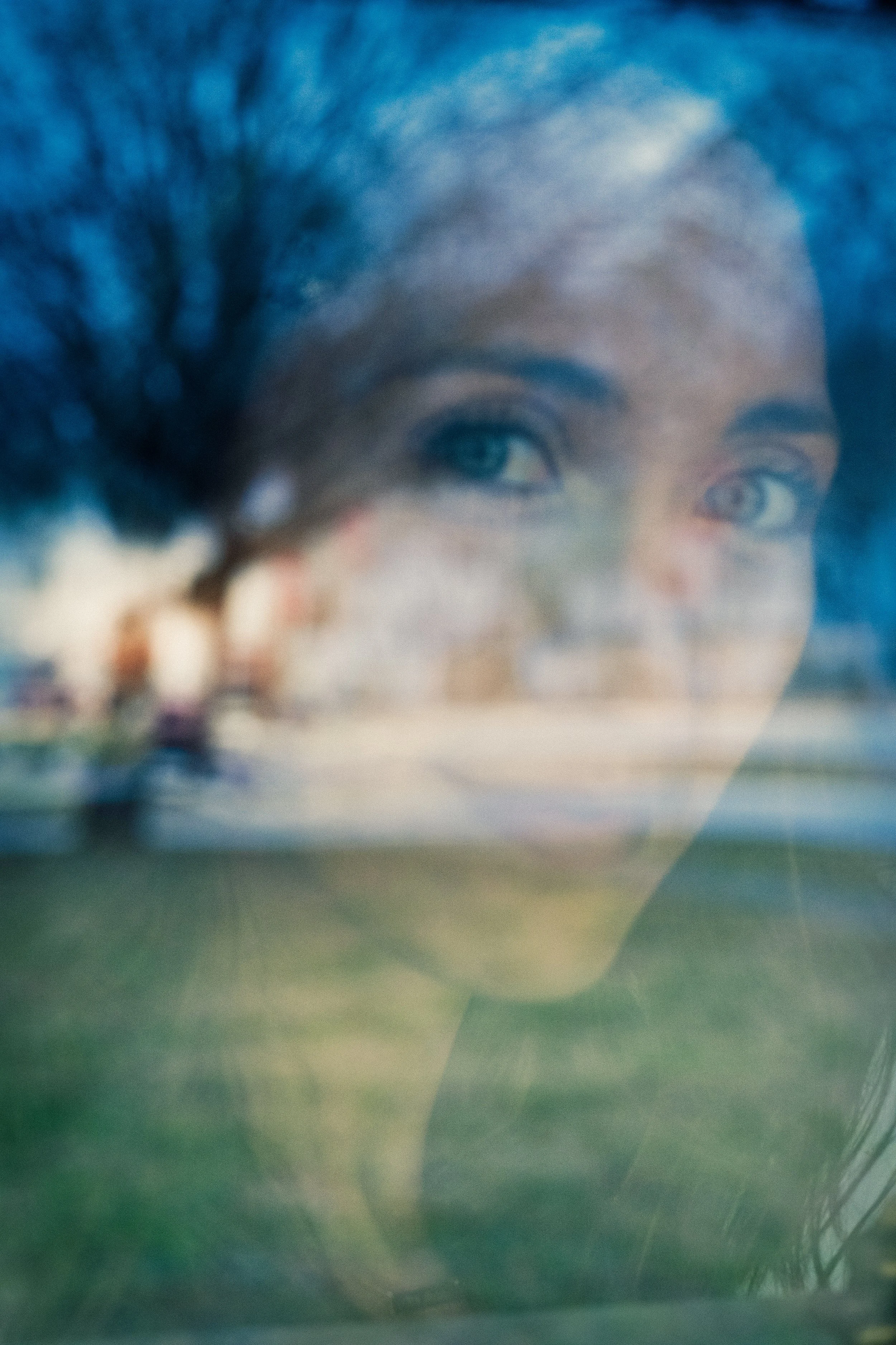 A double exposure photograph showing a woman's face reflected in a window with a blurred outdoor scene of trees and a park.
