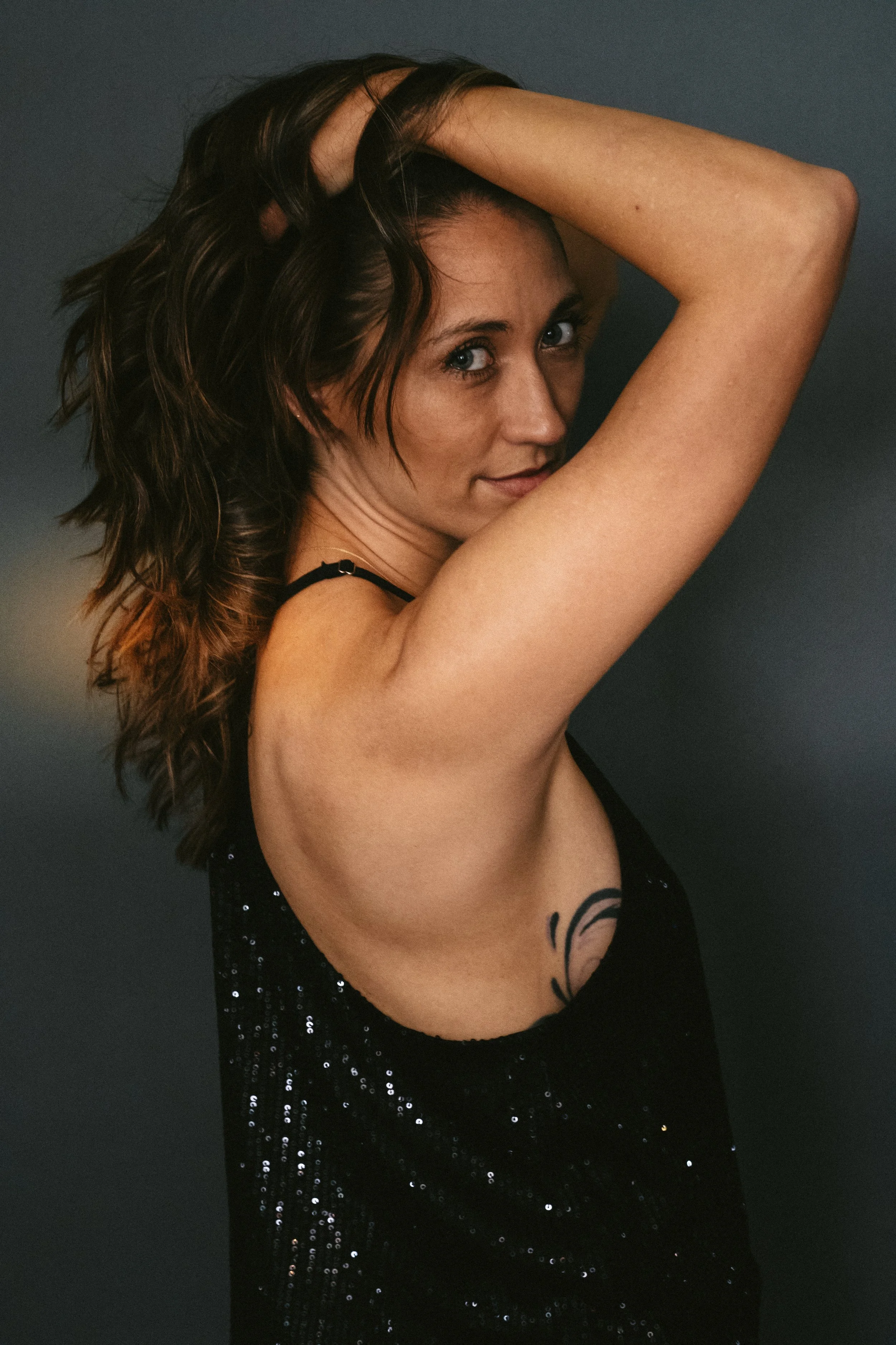 A woman with wavy brown hair and blue eyes, wearing a black sleeveless top with sequins, poses with one arm raised above her head, partially covering her hair, against a dark background.