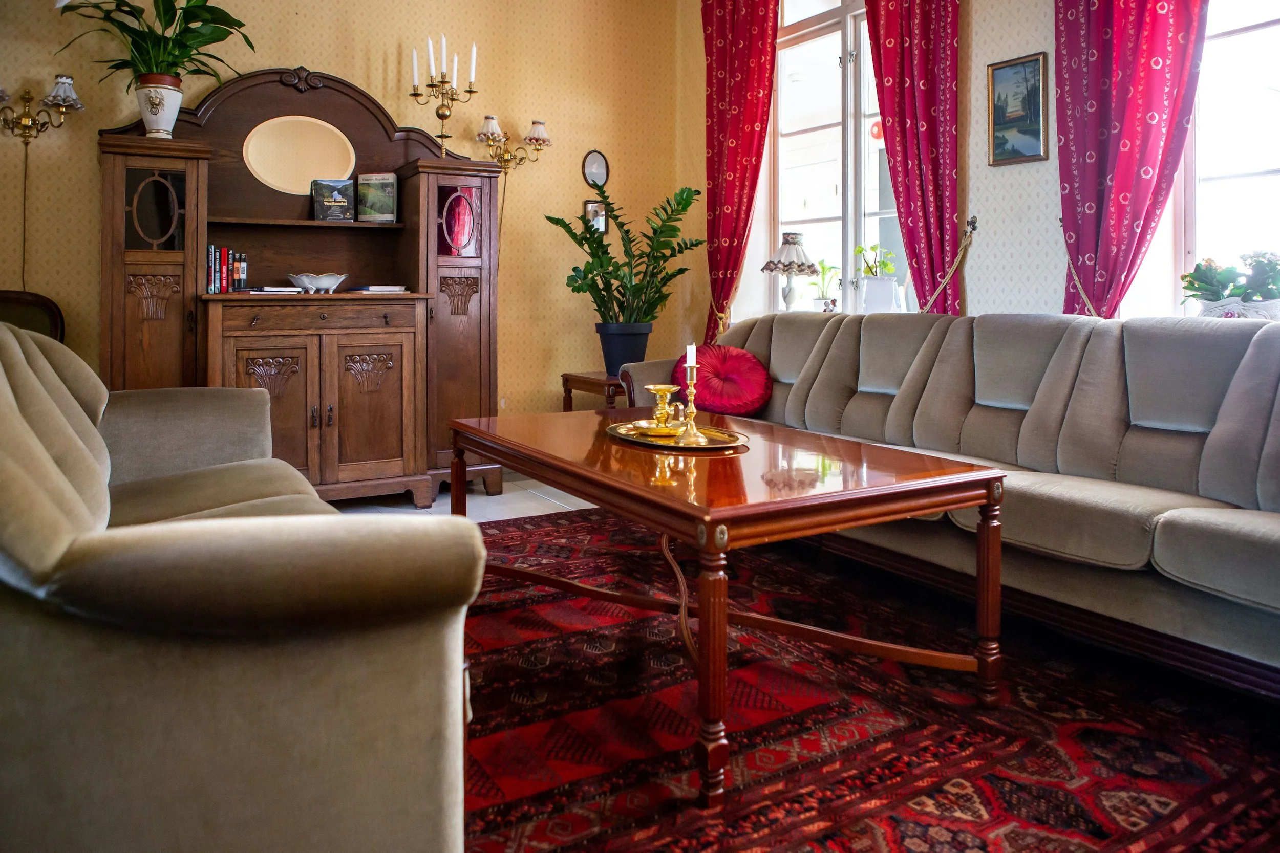 Et koselig stue med beige sofa og stol, et trebord med lysestaker, en stor plante, permer og bøker, store vinduer med røde gardiner, og en veggdekorasjon.