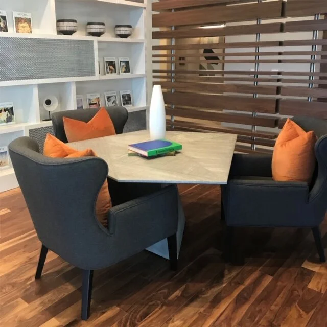 A cozy dining area with a marble table surrounded by four upholstered dark gray chairs with orange pillows. On the table are a tall white vase and a few stacked books. In the background, a white shelving unit holds framed photos, books, and decorative bowls, and a wooden slat partition separates the space from another room.