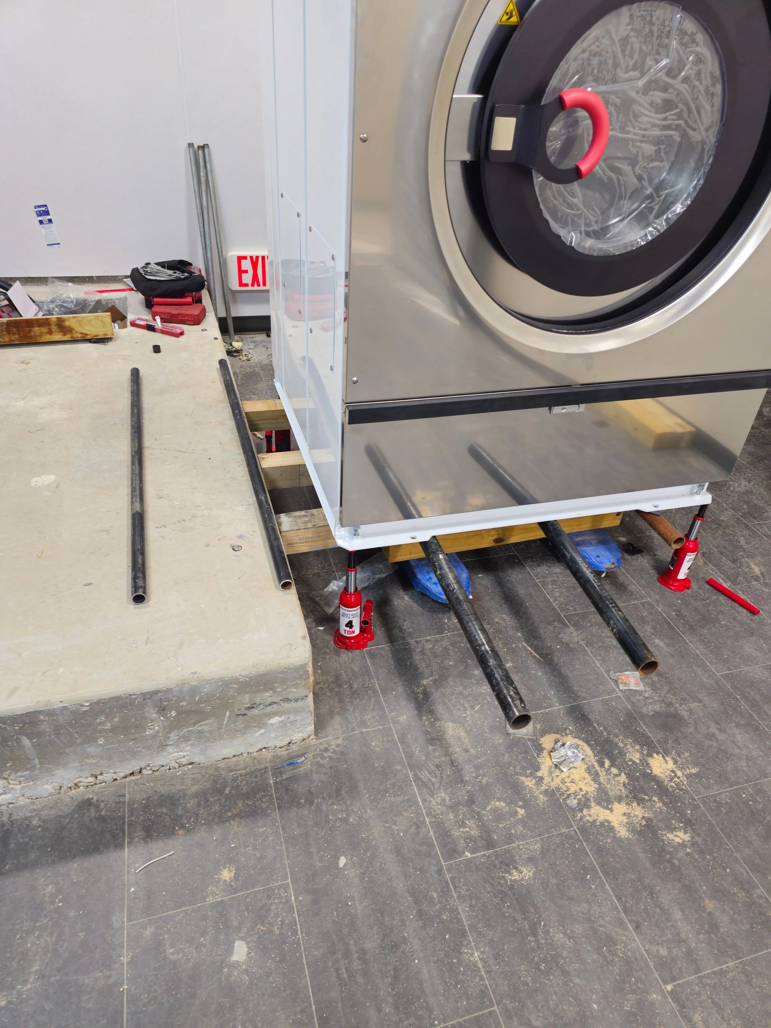 Construction site with a large commercial dryer being installed. Metal pipes and tools are scattered on the floor, with red jacks supporting the appliance. A concrete step is visible on the left, and an exit sign is in the background.