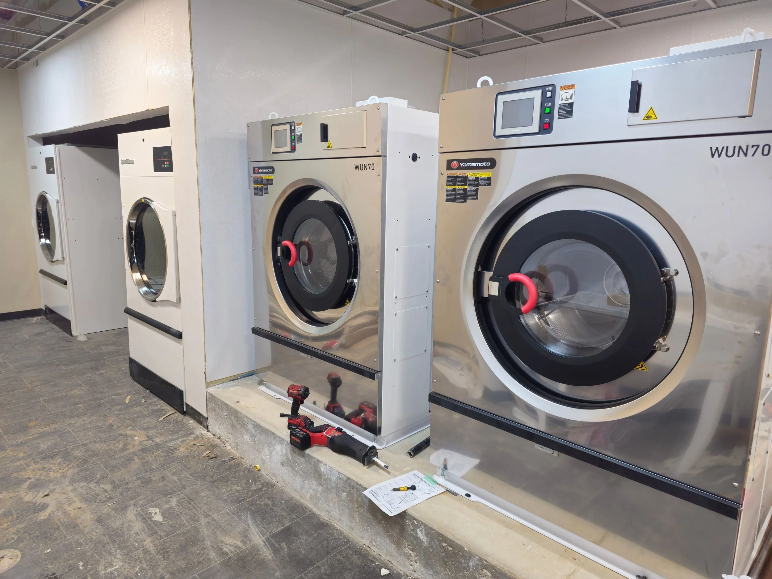 A row of commercial laundry washing machines in a laundry facility, with tools and instruction manual on the floor in front of them.