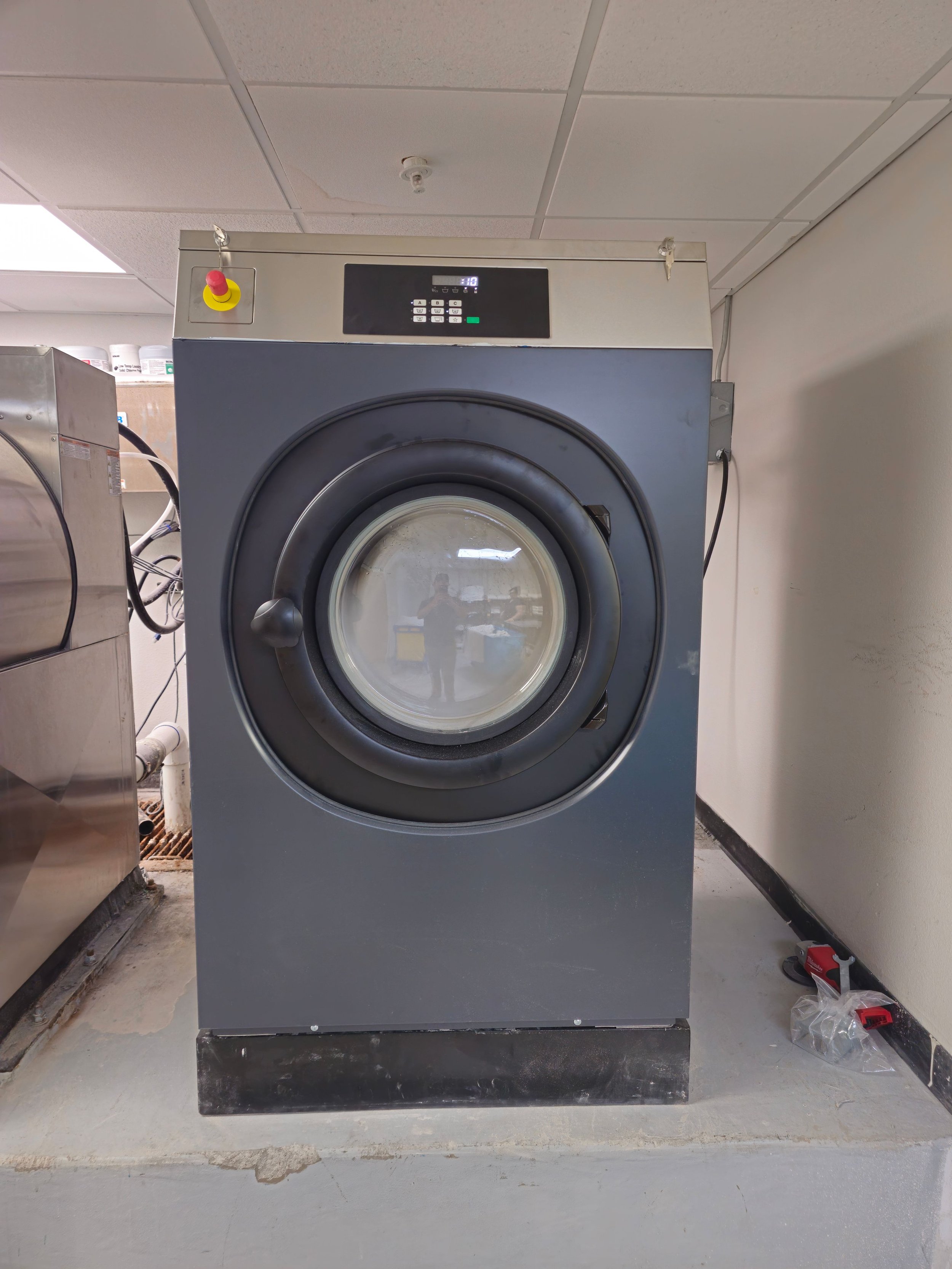 Front view of a commercial washing machine in a laundry room, with a glass door showing the inner drum.