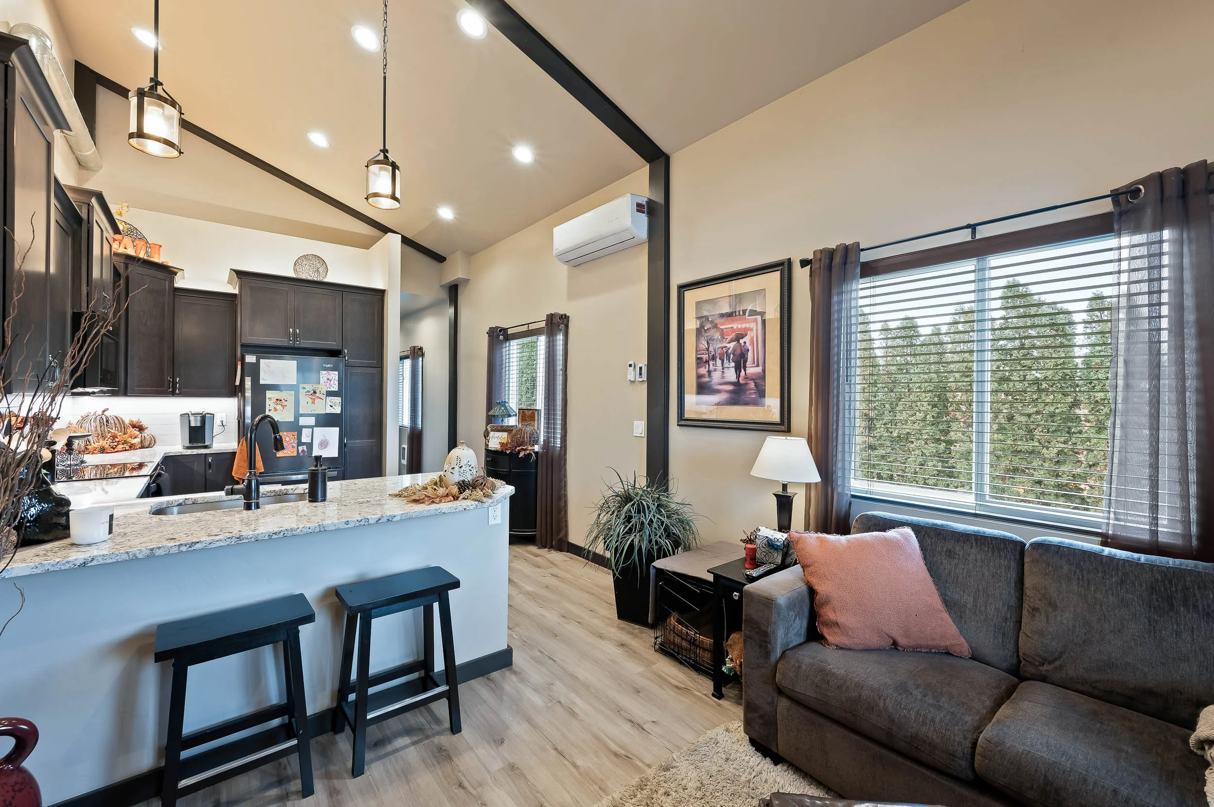 Open-concept living room and kitchen with dark wood cabinets, granite countertops, a gray couch with pillows, large windows with blinds, and autumn decorations.