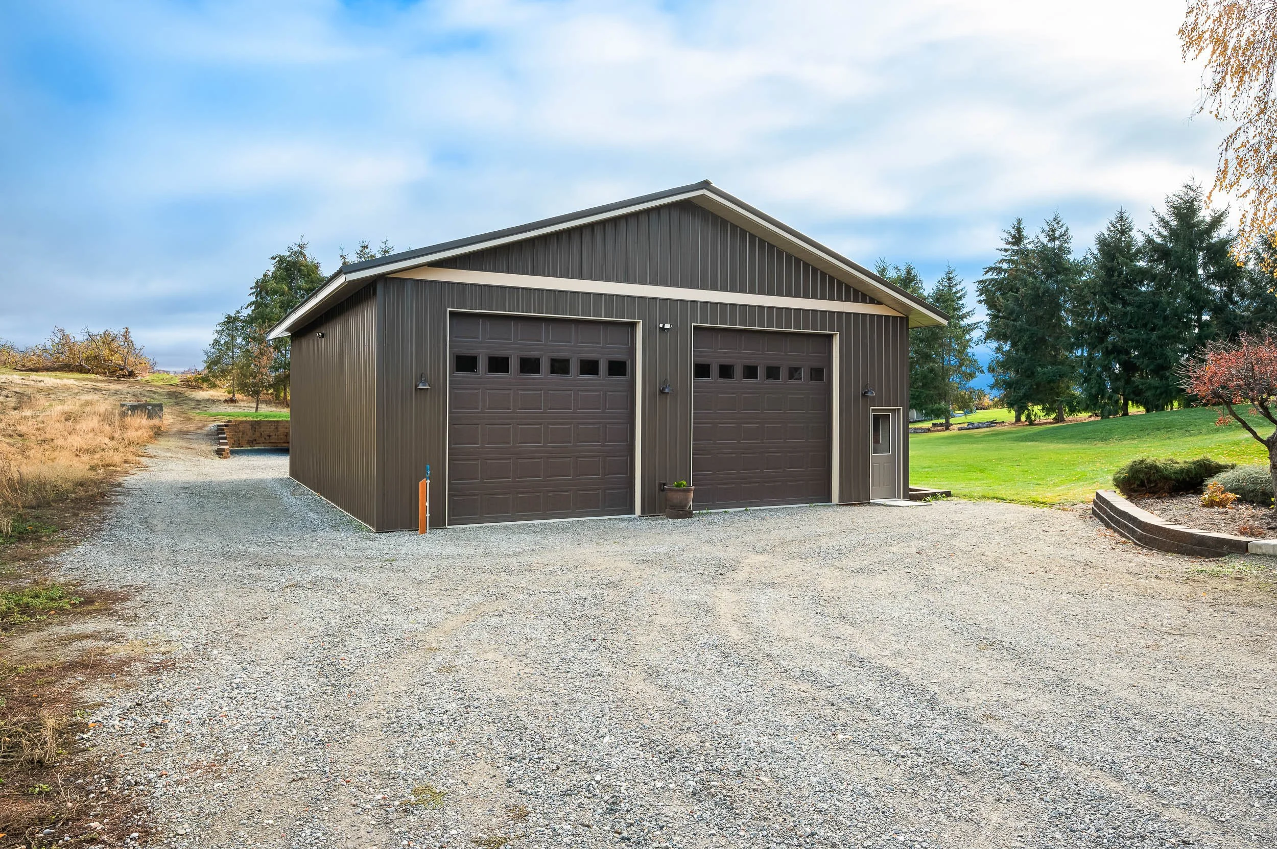 Large gray metal garage with two roller doors and a side door, situated on a gravel driveway surrounded by a grassy yard with trees.