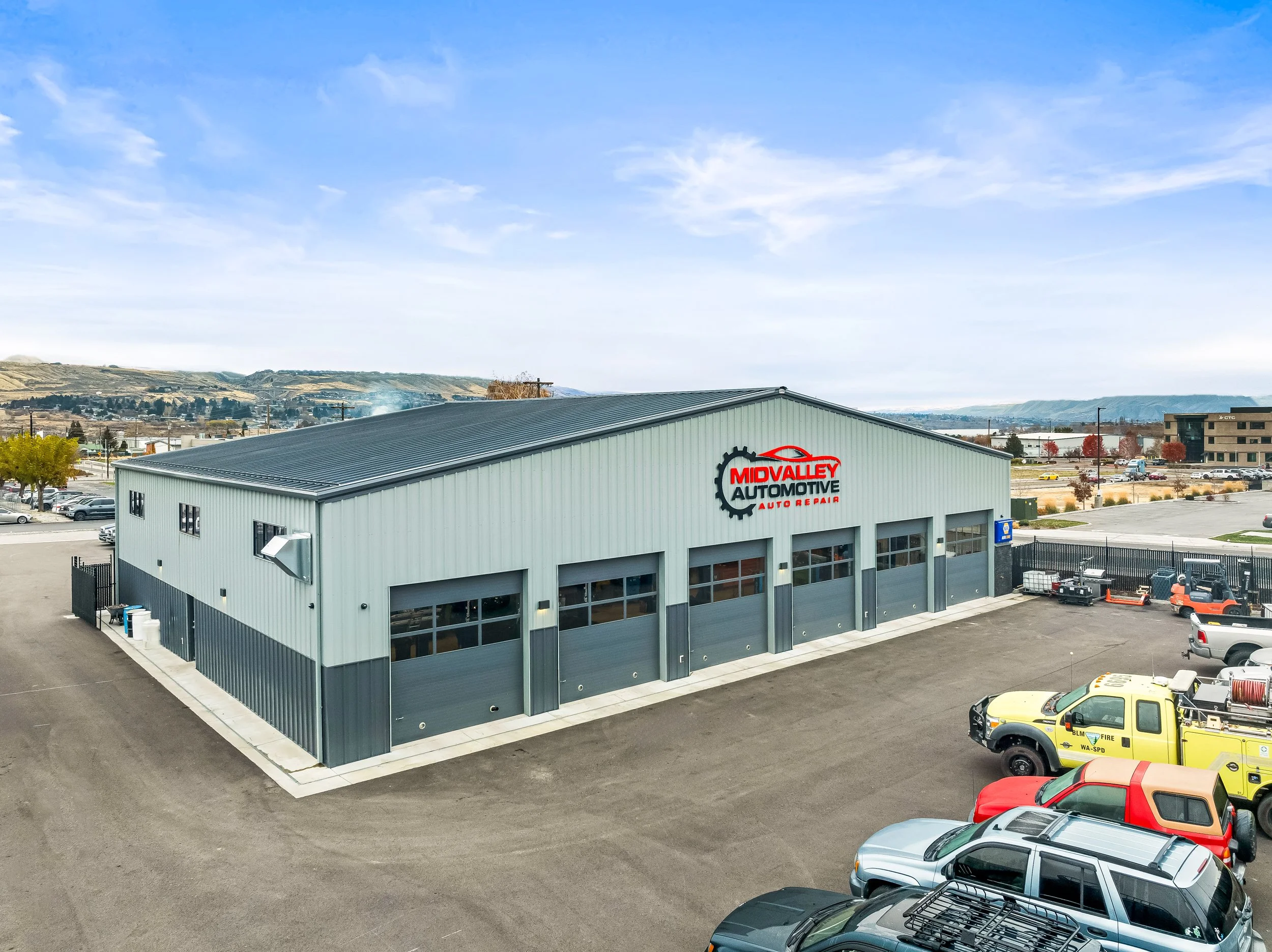 Automotive repair shop called Midvalley Automotive, a large gray metal building with four overhead garage doors and a sign displaying the business name and logo, surrounded by parking lot and vehicles including a yellow fire truck, with hills and a p
