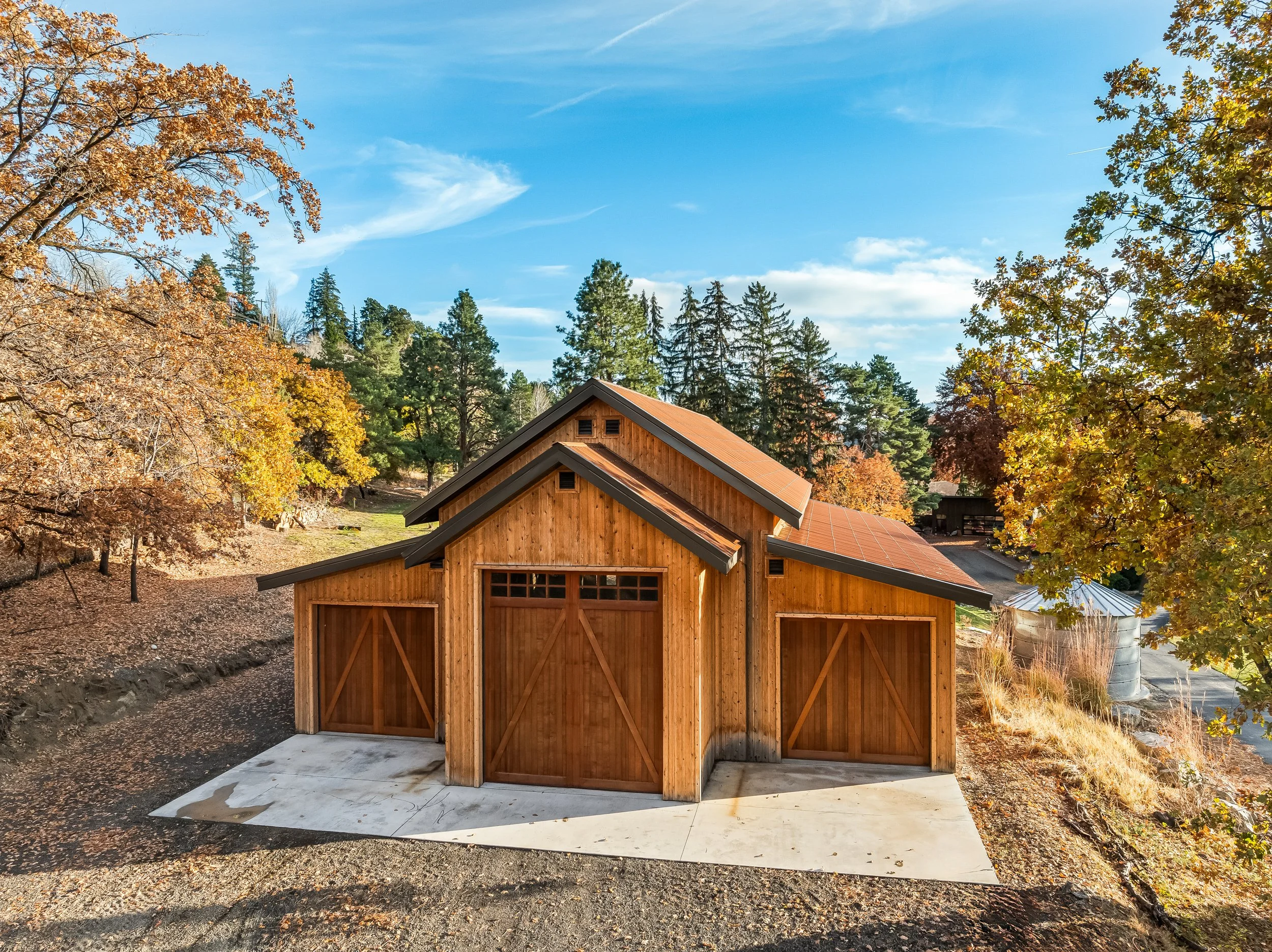 A wooden garage with two large doors, situated in a rural, wooded area during fall with colorful trees and a blue sky.