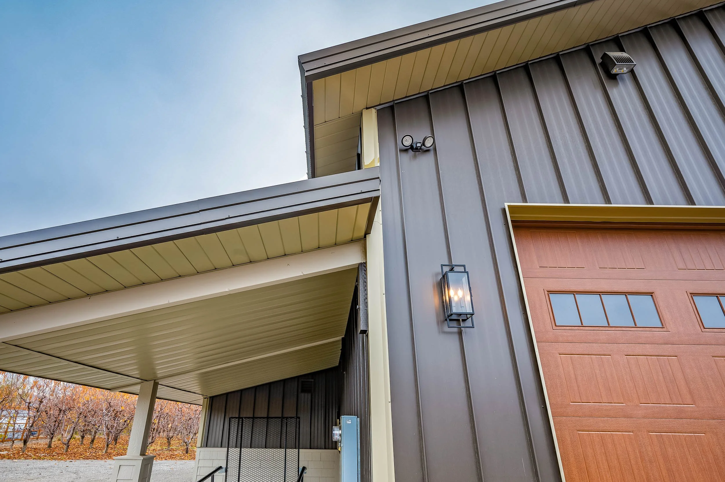 Close-up of a modern barn-style building with metal siding, a wooden garage door, outdoor wall lantern, security camera, and an understory porch overhang. In the background, a fruit orchard with leafless trees is visible.