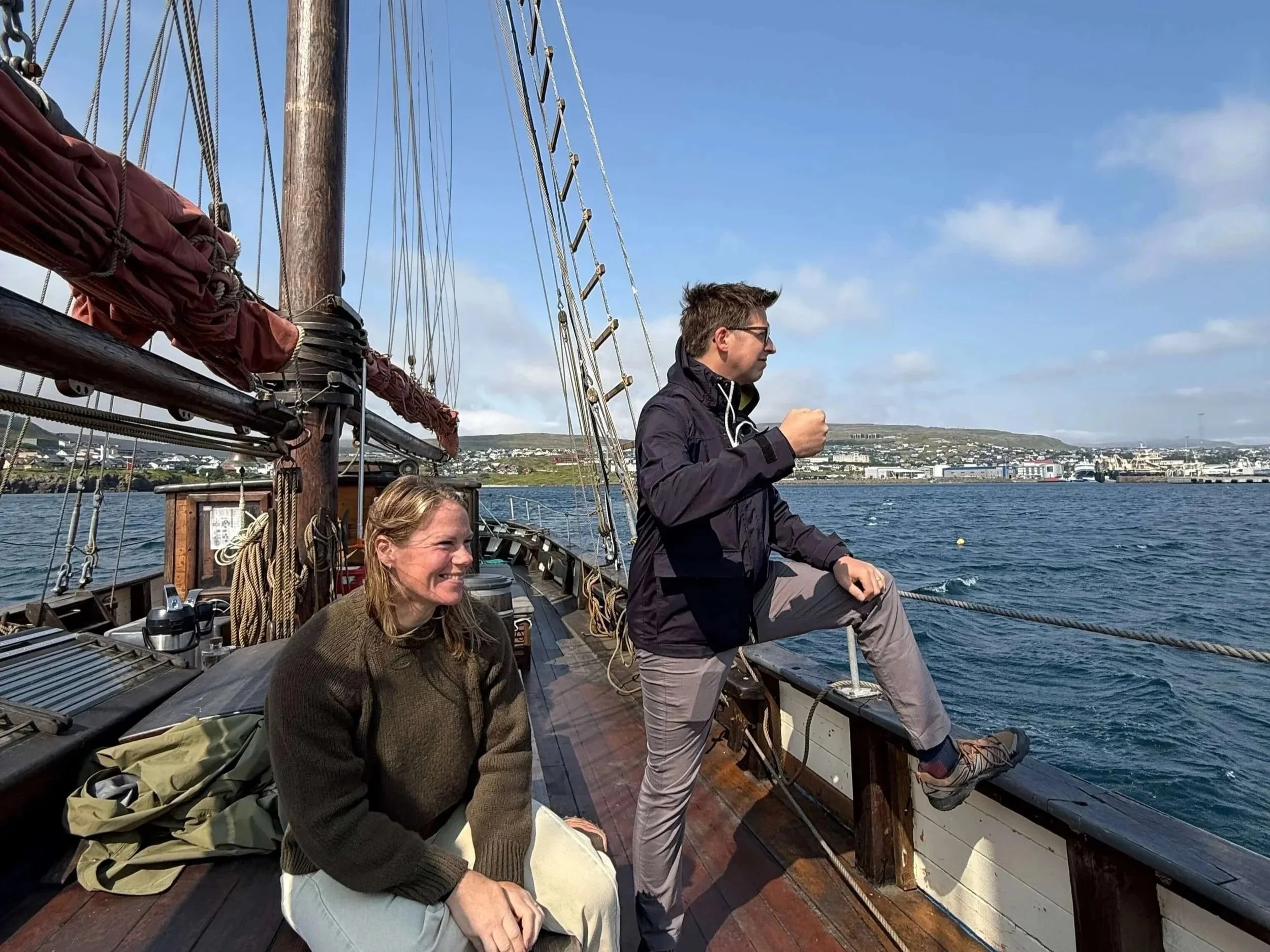 Three people on a sailboat, with two women sitting and a man standing, looking out at a cityscape across the water. The sailboat has ropes, wood, and rigging visible. The sky is partly cloudy.