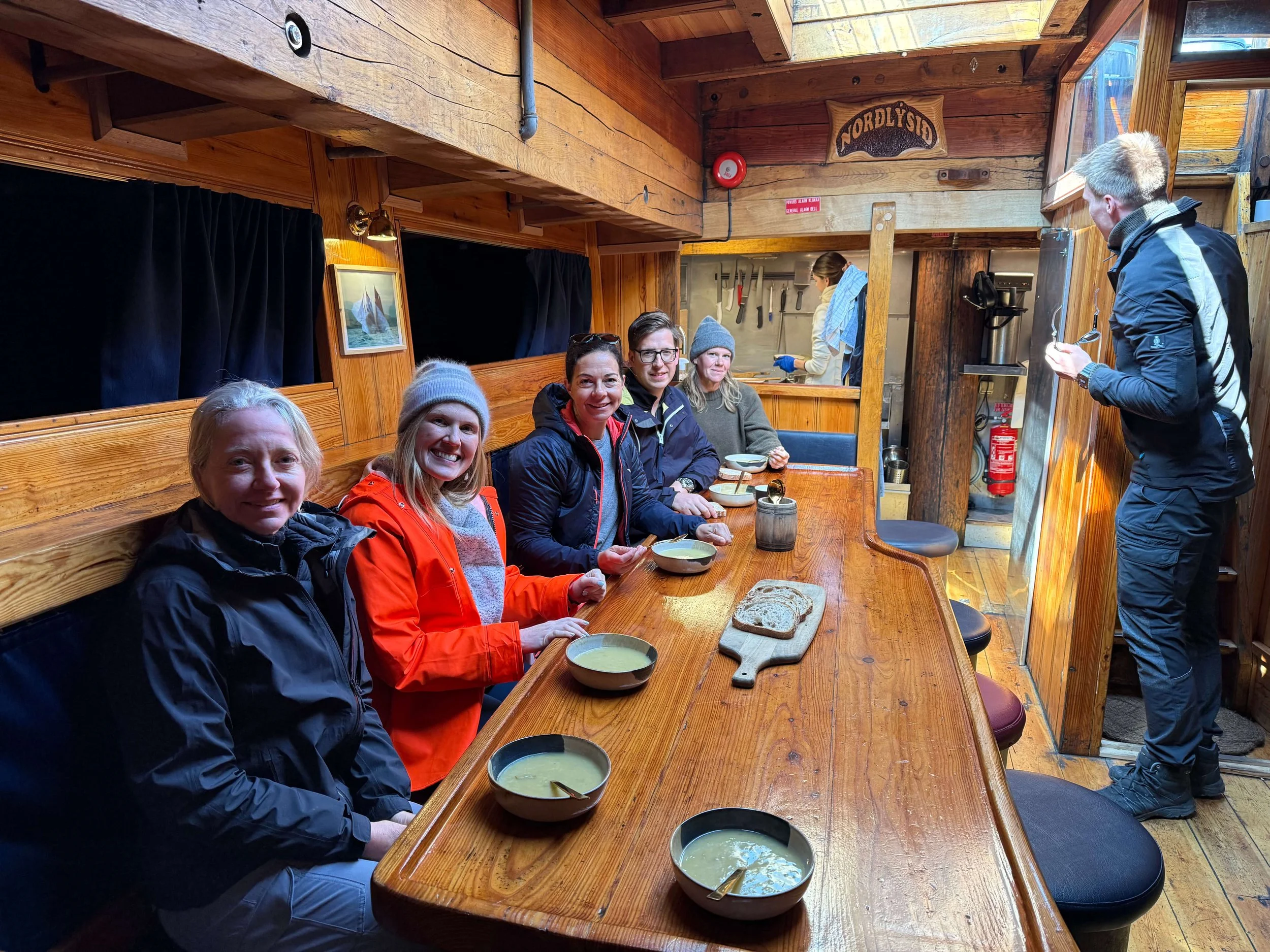 A group of four people sitting at a wooden counter inside a rustic restaurant, eating bowls of green soup. Behind them, a woman prepares food in the kitchen, which is visible through an open window. The interior features wooden walls and a sign that reads 'Nordfjord'.