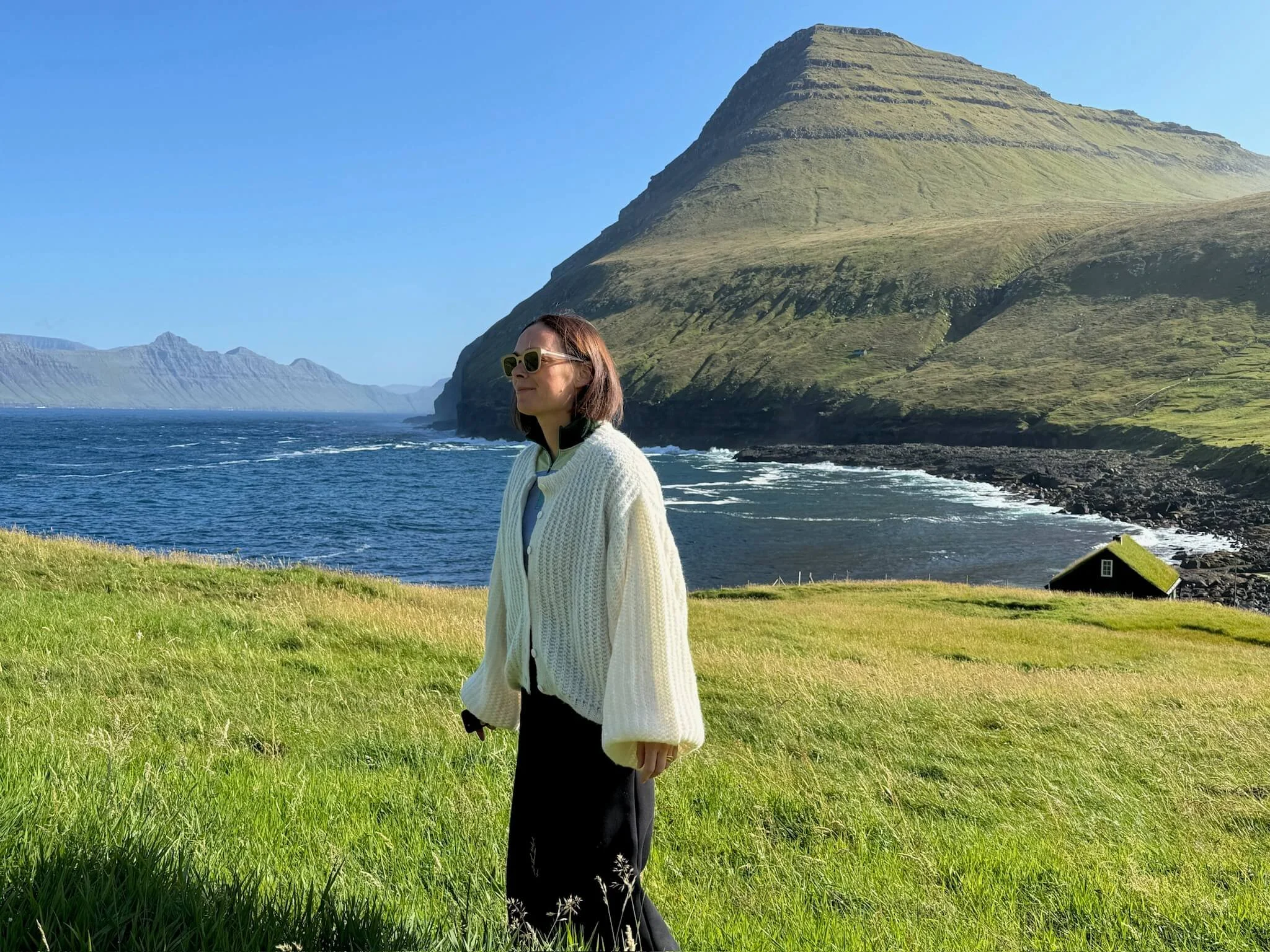 A woman wearing sunglasses and a white cardigan stands in a grassy field with a scenic coastal landscape, including cliffs and a house, in the background.