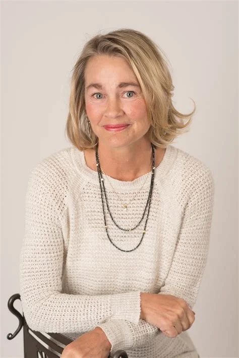 A woman with shoulder-length blonde hair, wearing a white knitted sweater and layered necklaces, sitting against a plain light-colored background.