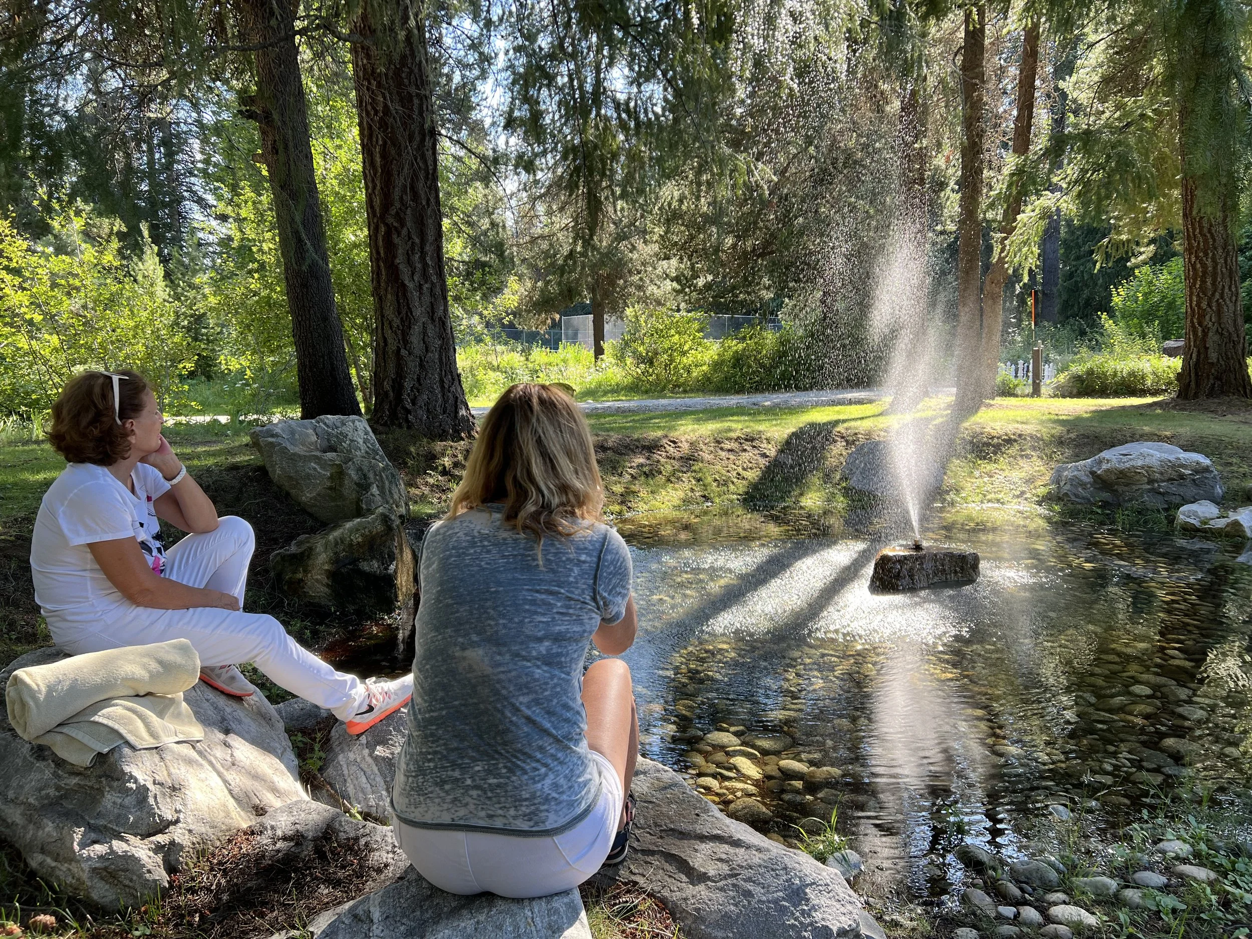 Two women sitting on rocks near a small pond, one woman is facing away, and the other two are facing each other, with trees and foliage in the background. Water from a fountain sprays into the pond.
