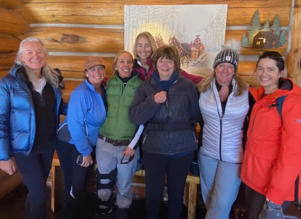 Group of seven women in outdoor winter clothing smiling in a log cabin with wood-paneled walls and a winter scene painting behind them.