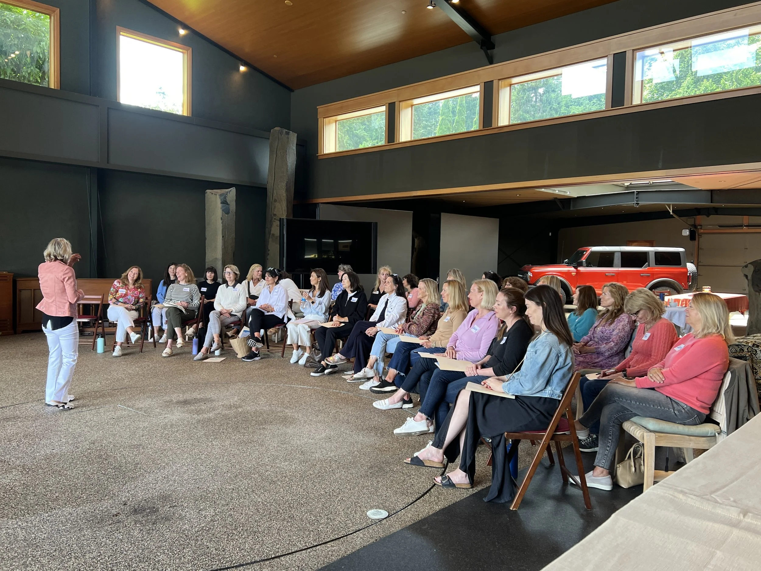 A group of women seated in rows attending a presentation in a spacious indoor venue with high ceilings, large windows, and a wooden ceiling. A woman in a pink blazer stands at the front speaking to the audience. There is a red off-road vehicle and large rock-like structures in the background.