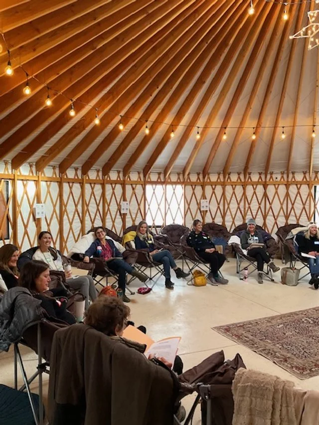 Group of people sitting in a circle inside a yurt, participating in a meeting or workshop, with warm lighting and cozy decor.