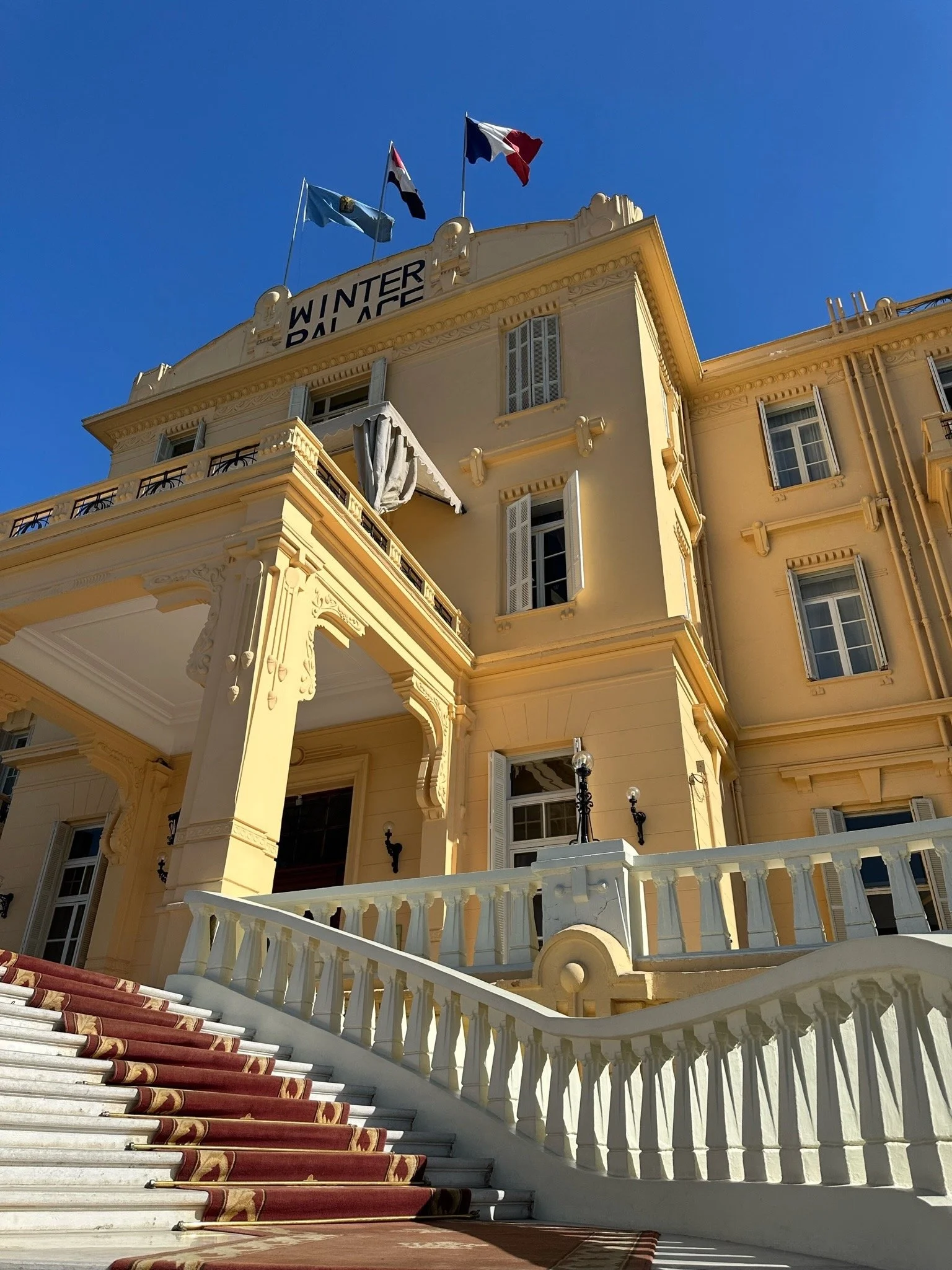 Exterior of a grand, yellow building named 'Winter Palace' with a staircase leading up to the entrance, flags on top (including the French flag), and a sign reading 'WINTER PALACE' at the top of the building.
