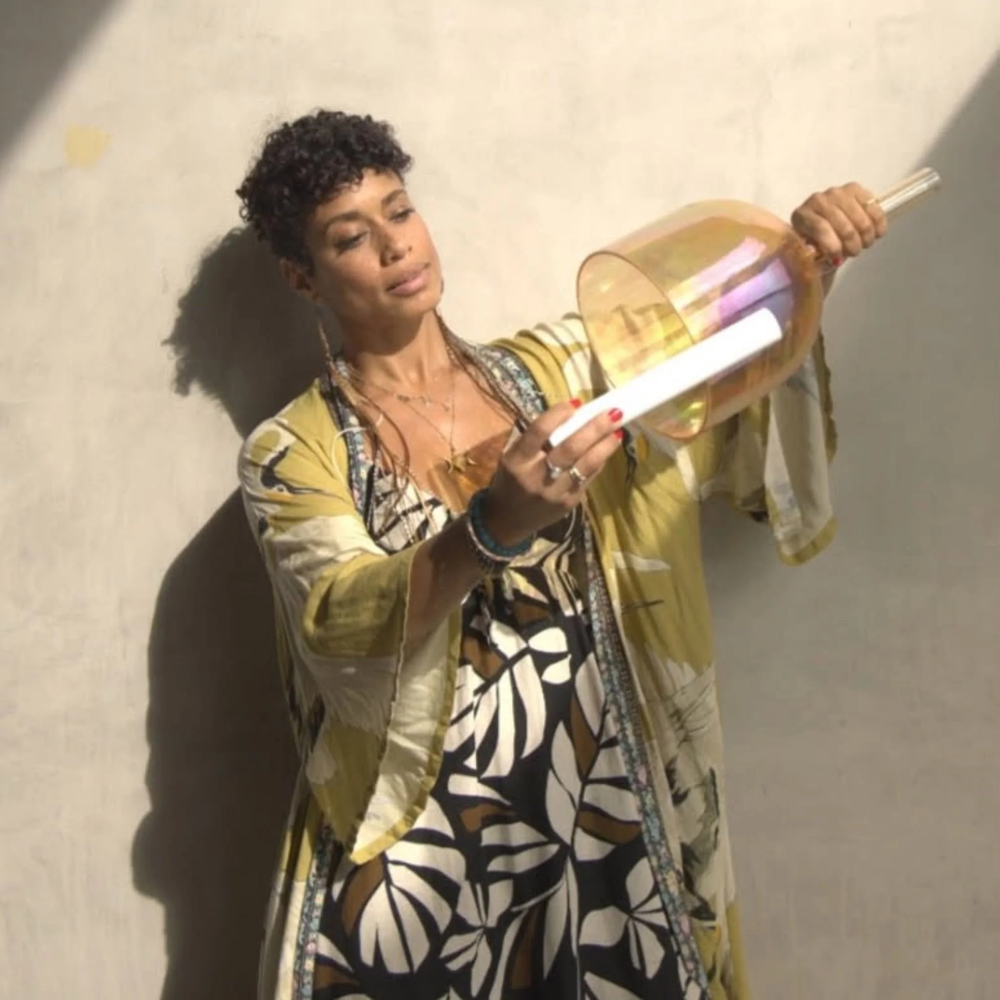 Woman with dark curly hair wearing a yellow and patterned dress, holding a large transparent instrument, standing against a plain wall.