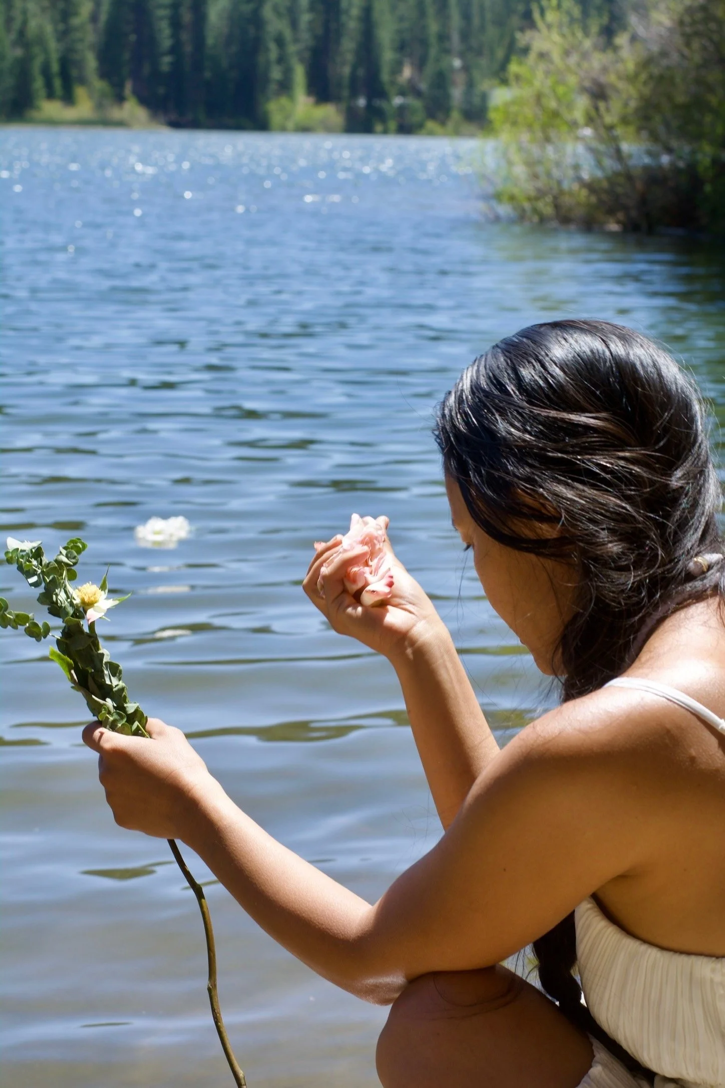 A woman sitting by a lake, holding a flower and a pink cloth, with trees in the background.