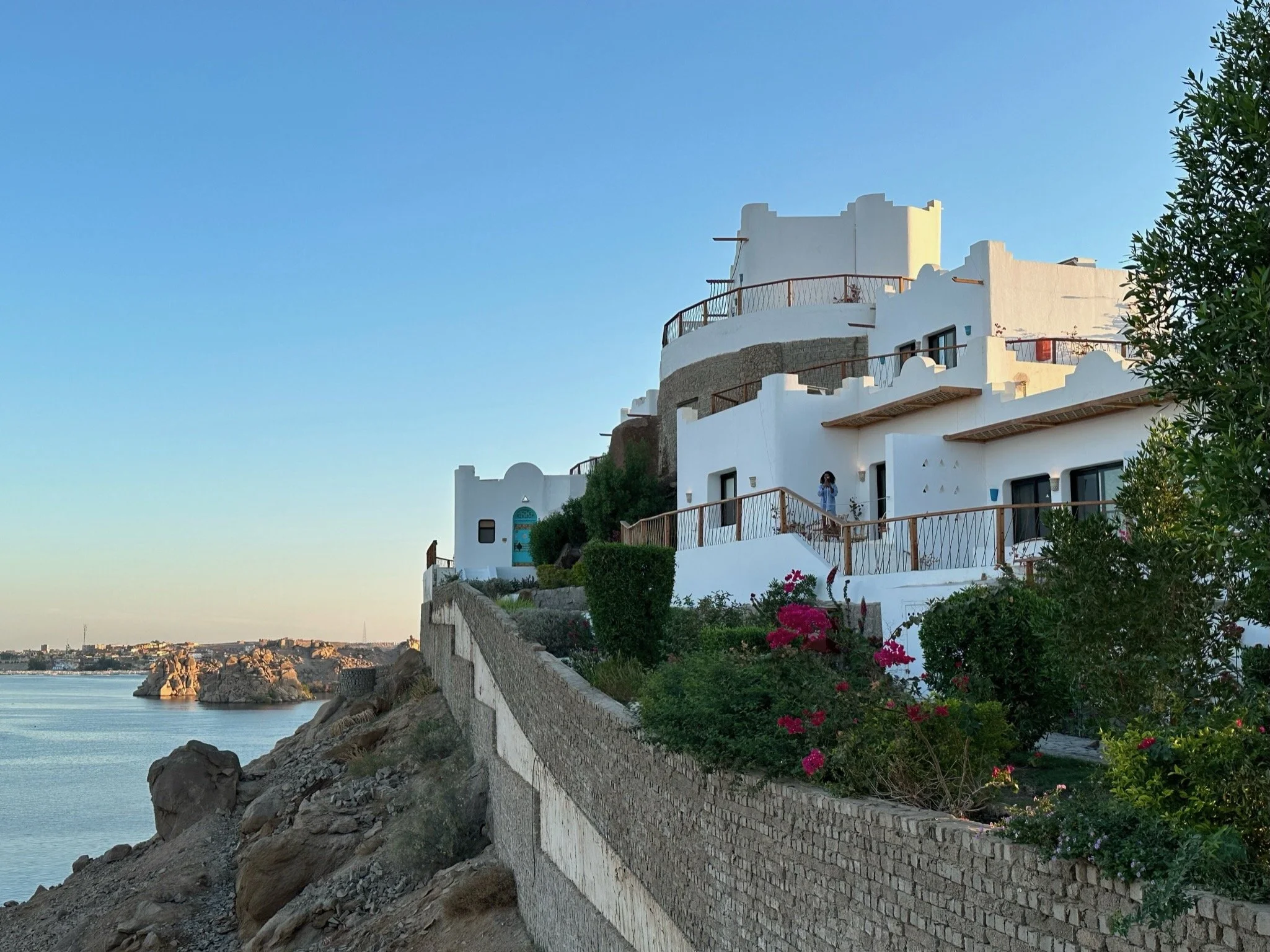 White Mediterranean-style building with multiple levels and balconies, situated on a rocky coastline with flowers and greenery, overlooking the ocean at sunset.