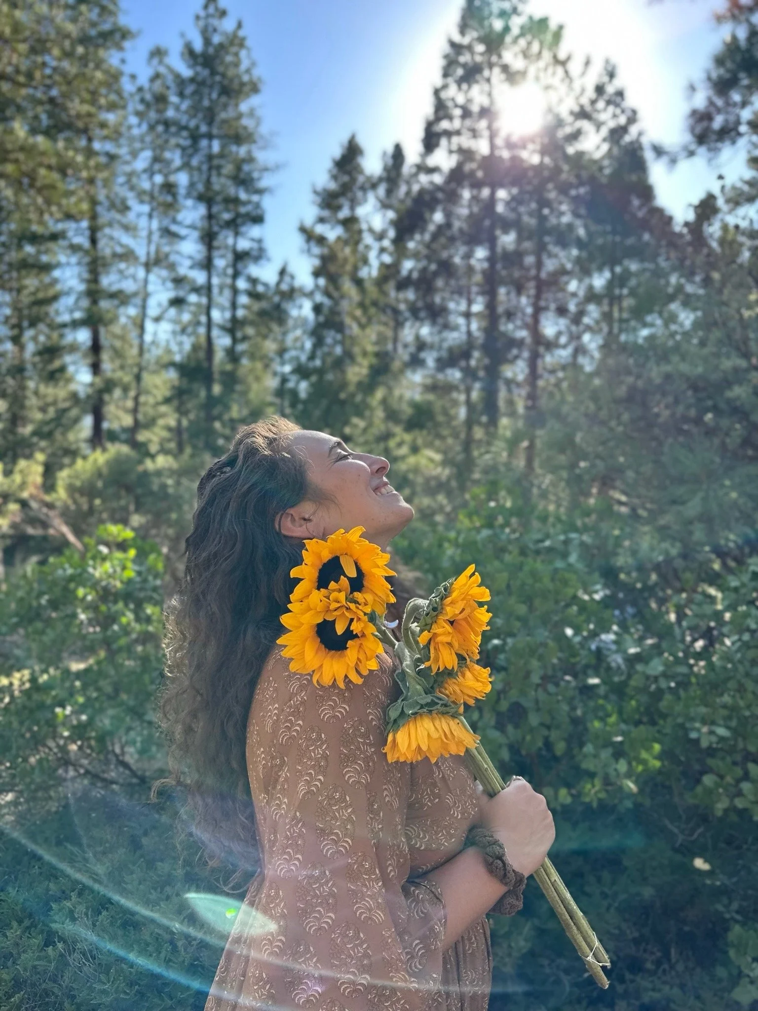 A woman smiling and enjoying sunlight in a forested area, holding a bouquet of sunflowers, wearing a patterned brown dress and a bracelet.