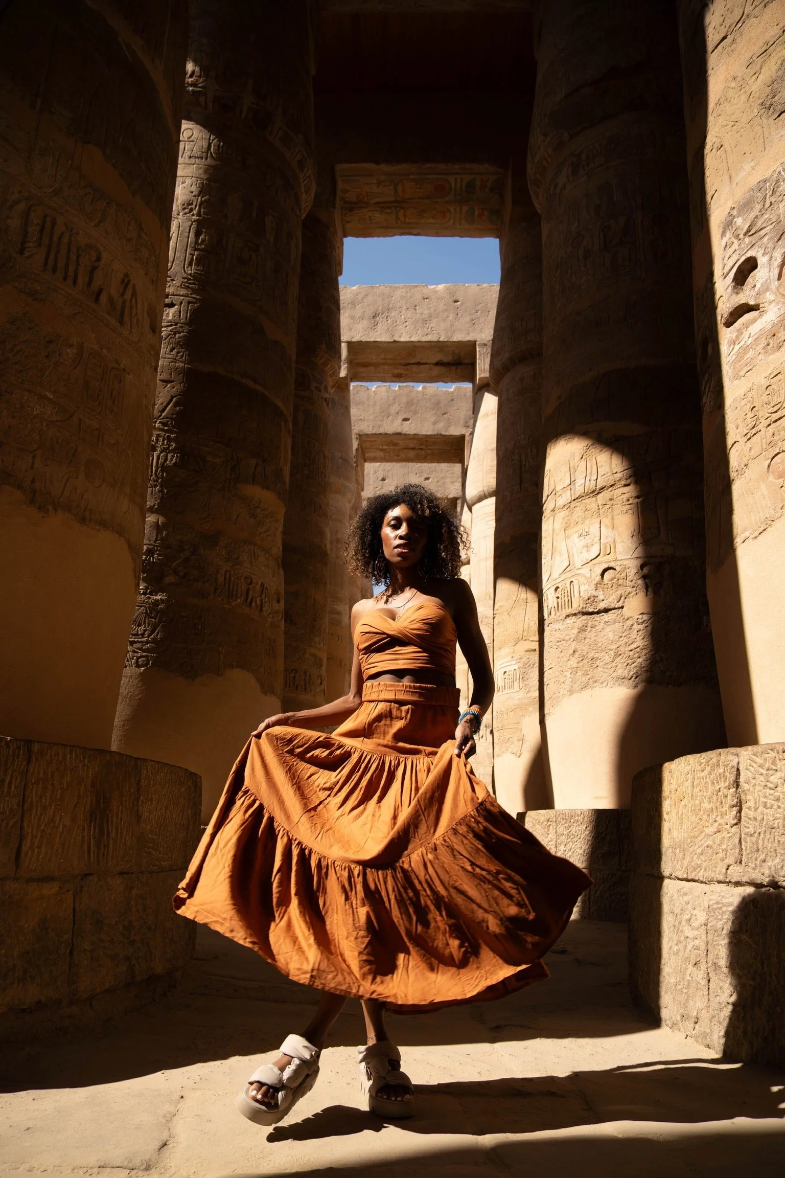 A woman wearing a brown skirt and crop top poses inside an ancient Egyptian temple with large stone columns covered in hieroglyphics, with a clear blue sky visible overhead.