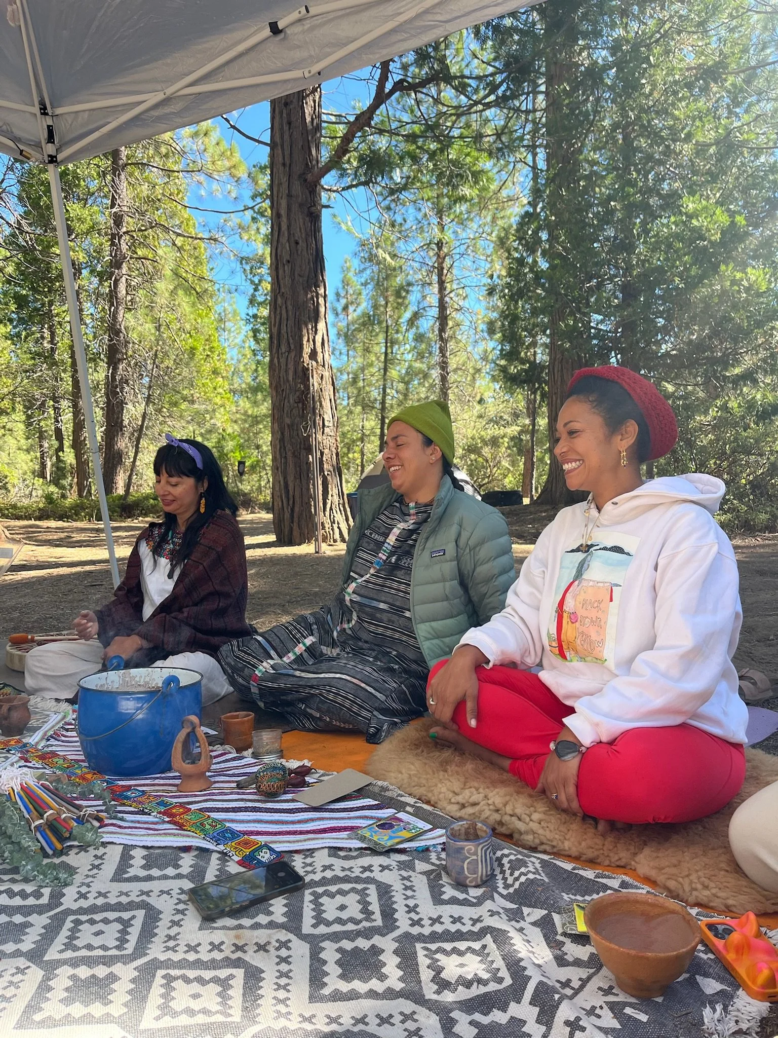 Three women sitting on blankets outdoors in a forest, smiling and laughing. They are surrounded by various items including bowls, candles, and a patterned cloth. They are dressed casually with colorful accessories.