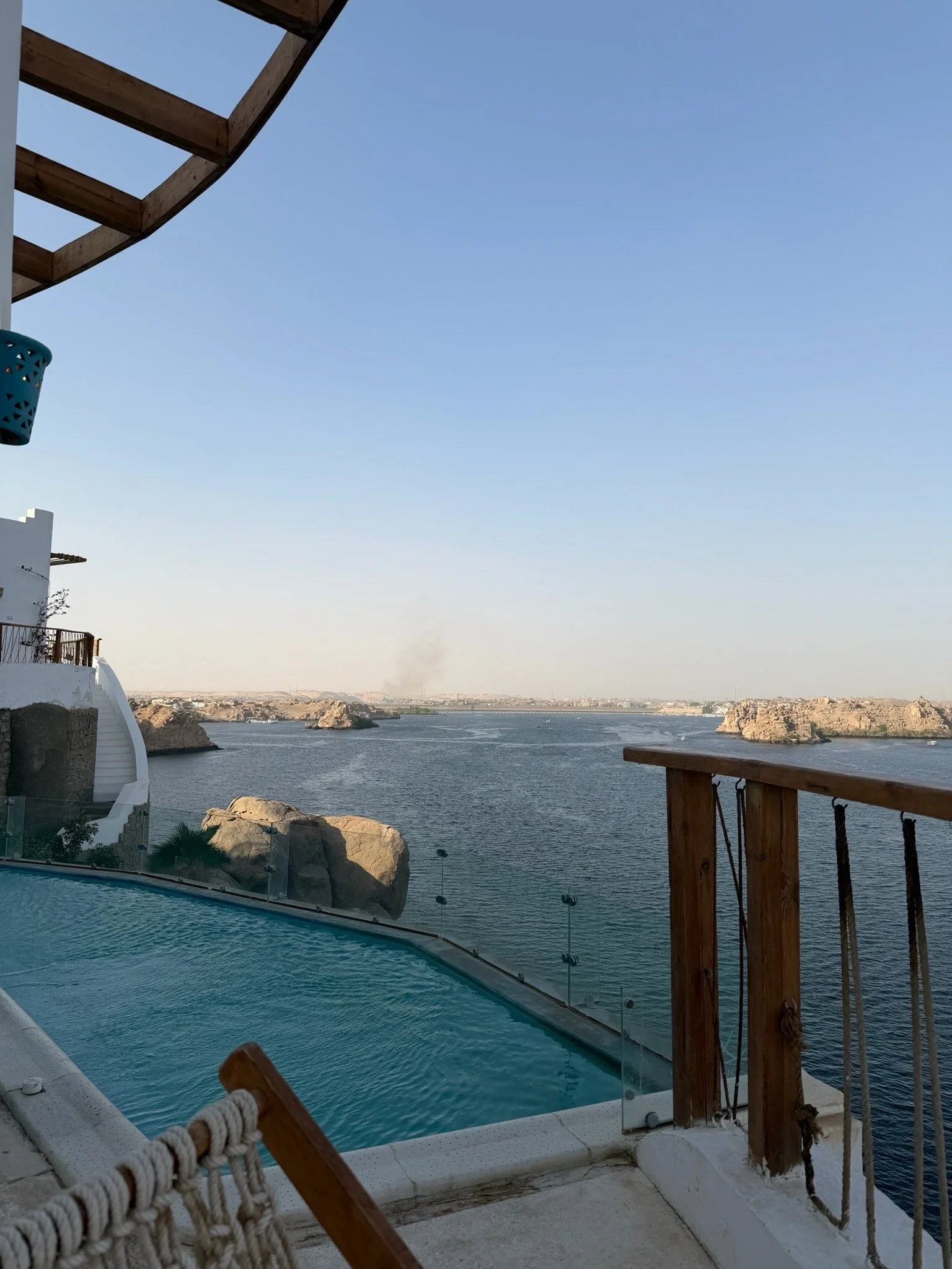 A view from a terrace overlooking a body of water with rocky islands and cliffs in the distance. Part of a wooden deck and a swimming pool are visible in the foreground.