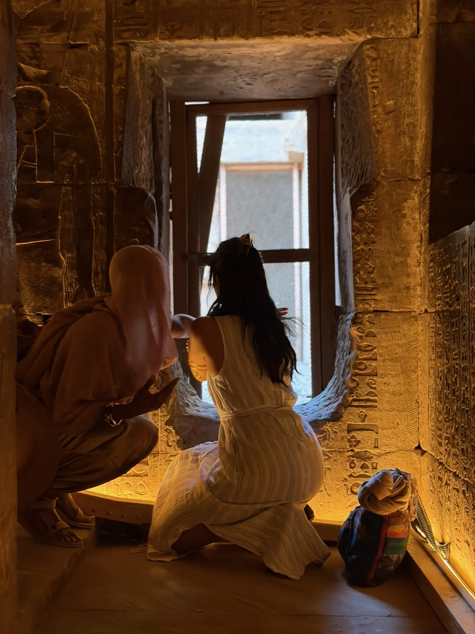 Two women on the retreat sitting and crouching near a window with ancient Egyptian hieroglyphs carved into stone walls, illuminated by warm lighting.