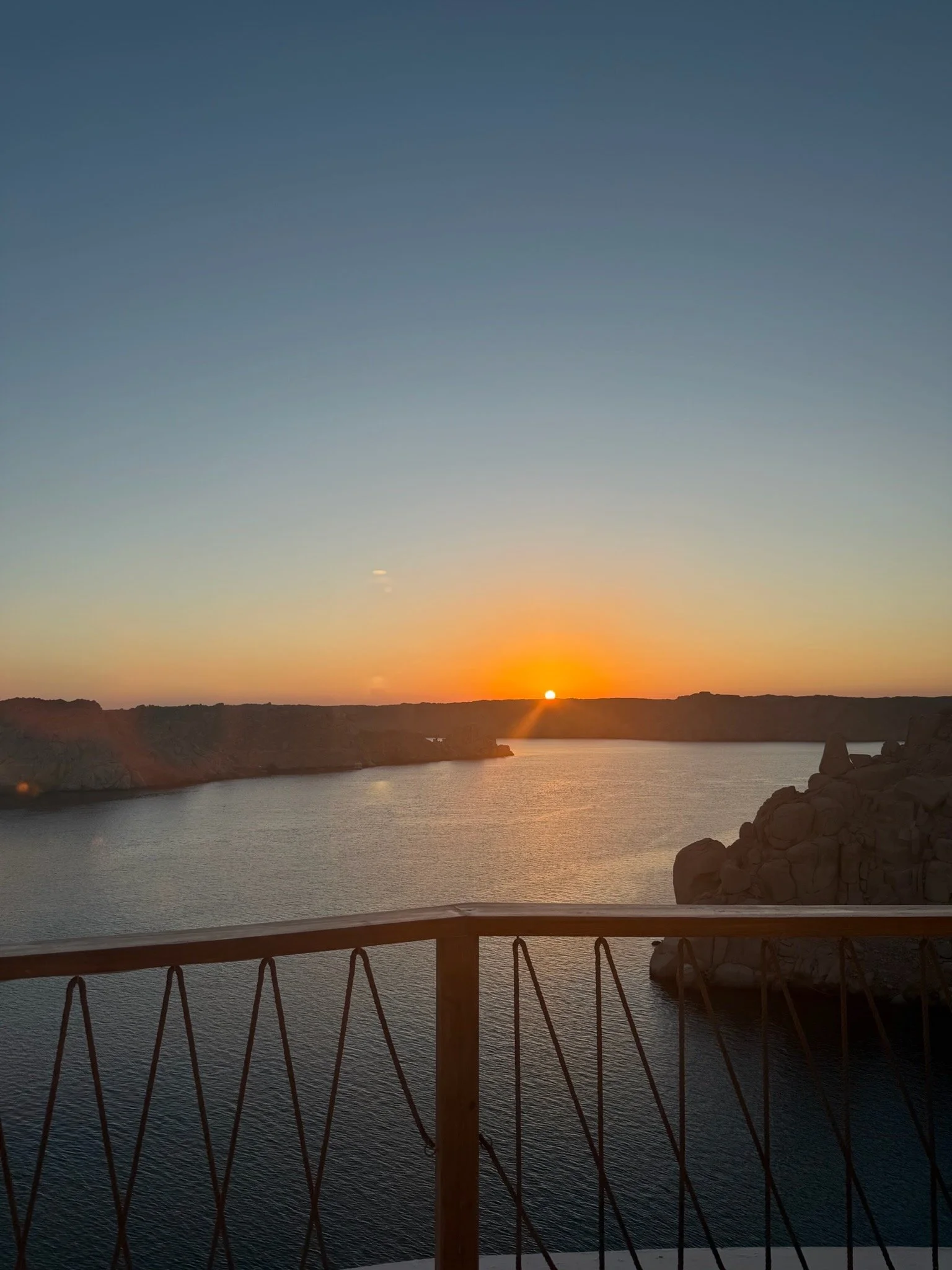 Sunset over a body of water, with rocky shoreline on the right and a wooden railing in the foreground.