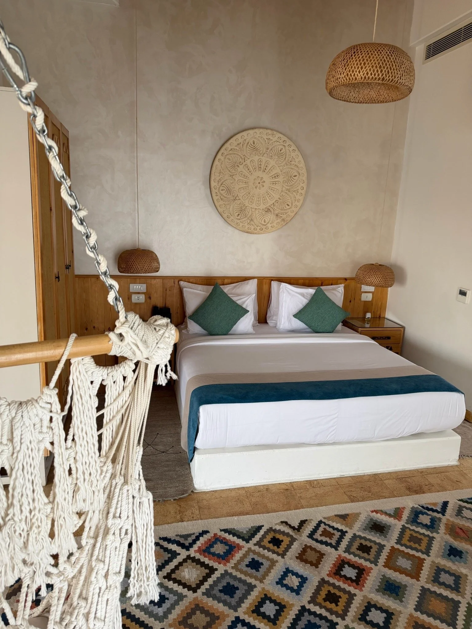 A hotel room with a bed, wooden headboard, green pillows, woven hanging lamps, a decorative wall piece, a colorful patterned rug, and a white hammock in the foreground.