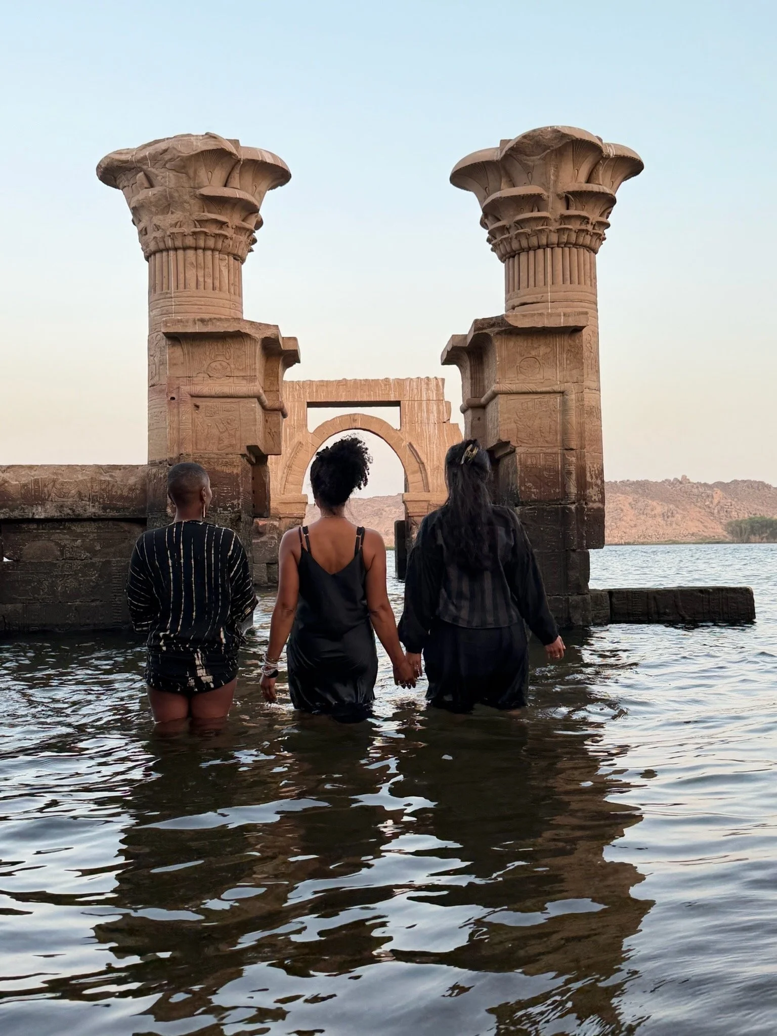 Three people in water holding hands, walking towards ancient Egyptian ruins with two large columns and an archway, overlooking a river or lake during sunset or sunrise.