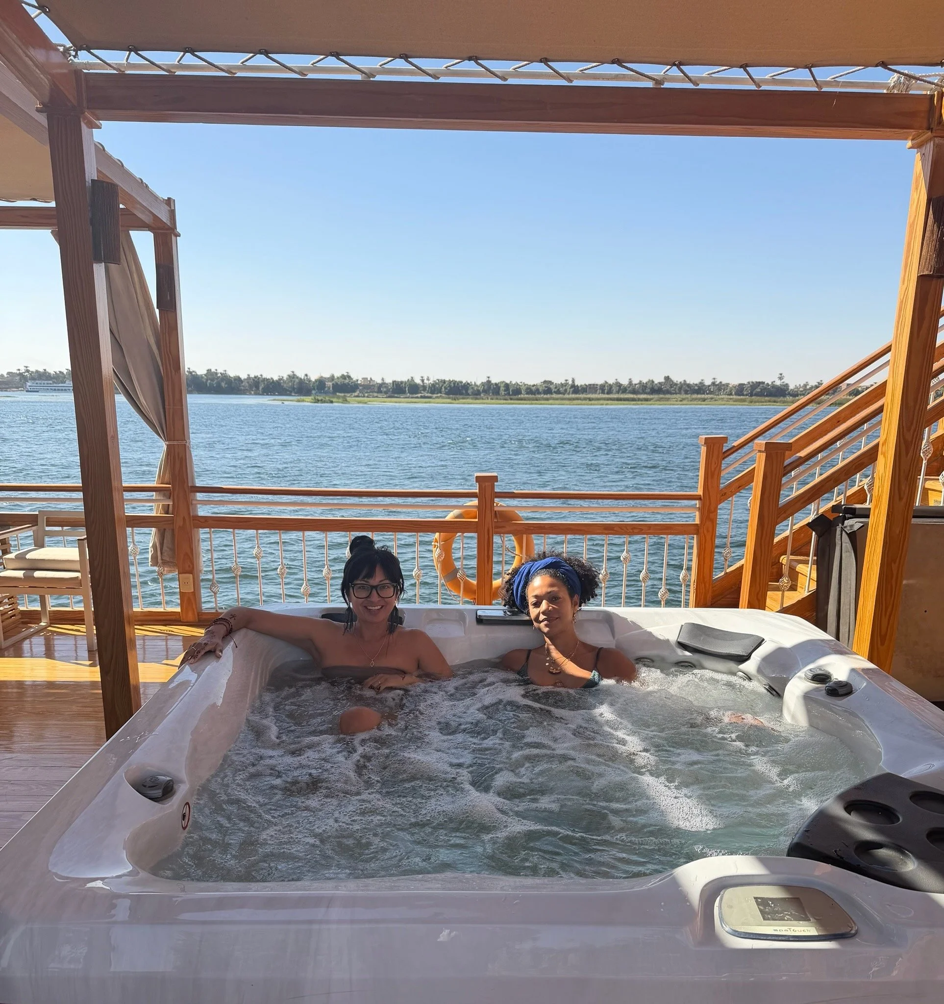 Two women relaxing in a hot tub on a boat with a large body of water and a distant shoreline in the background.