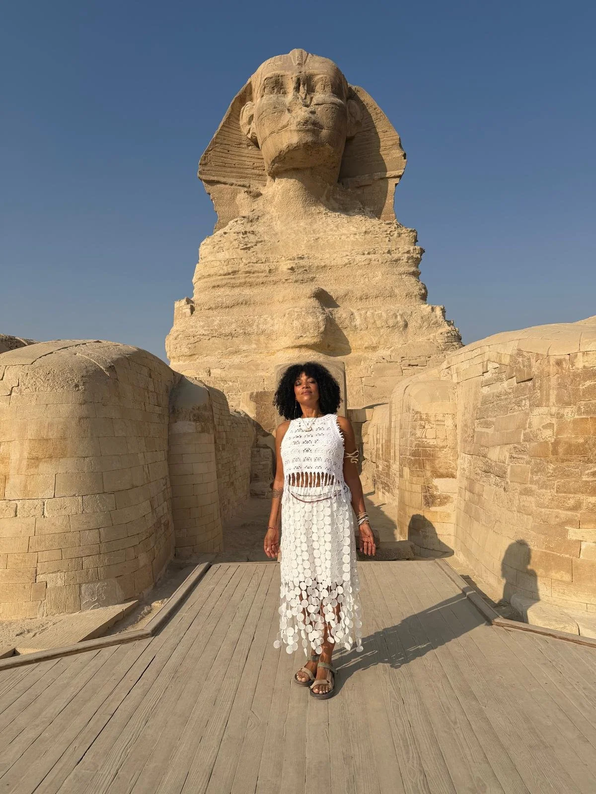 A woman standing on a wooden platform in front of the Great Sphinx of Giza during daytime, wearing a white crochet dress and sandals.