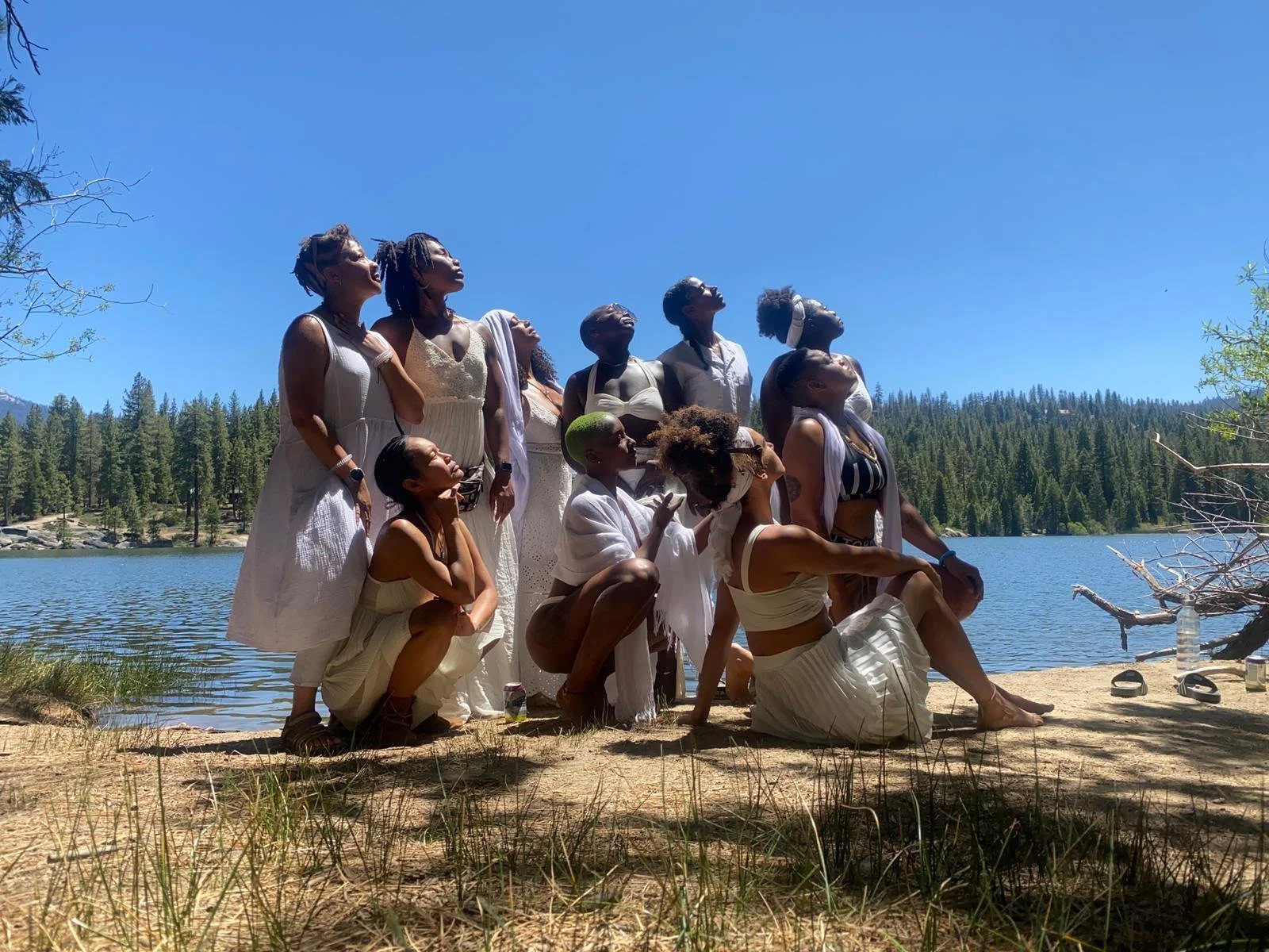 Group of people dressed in white clothing sitting and kneeling on sandy shore near a lake surrounded by pine trees, with clear blue sky above.