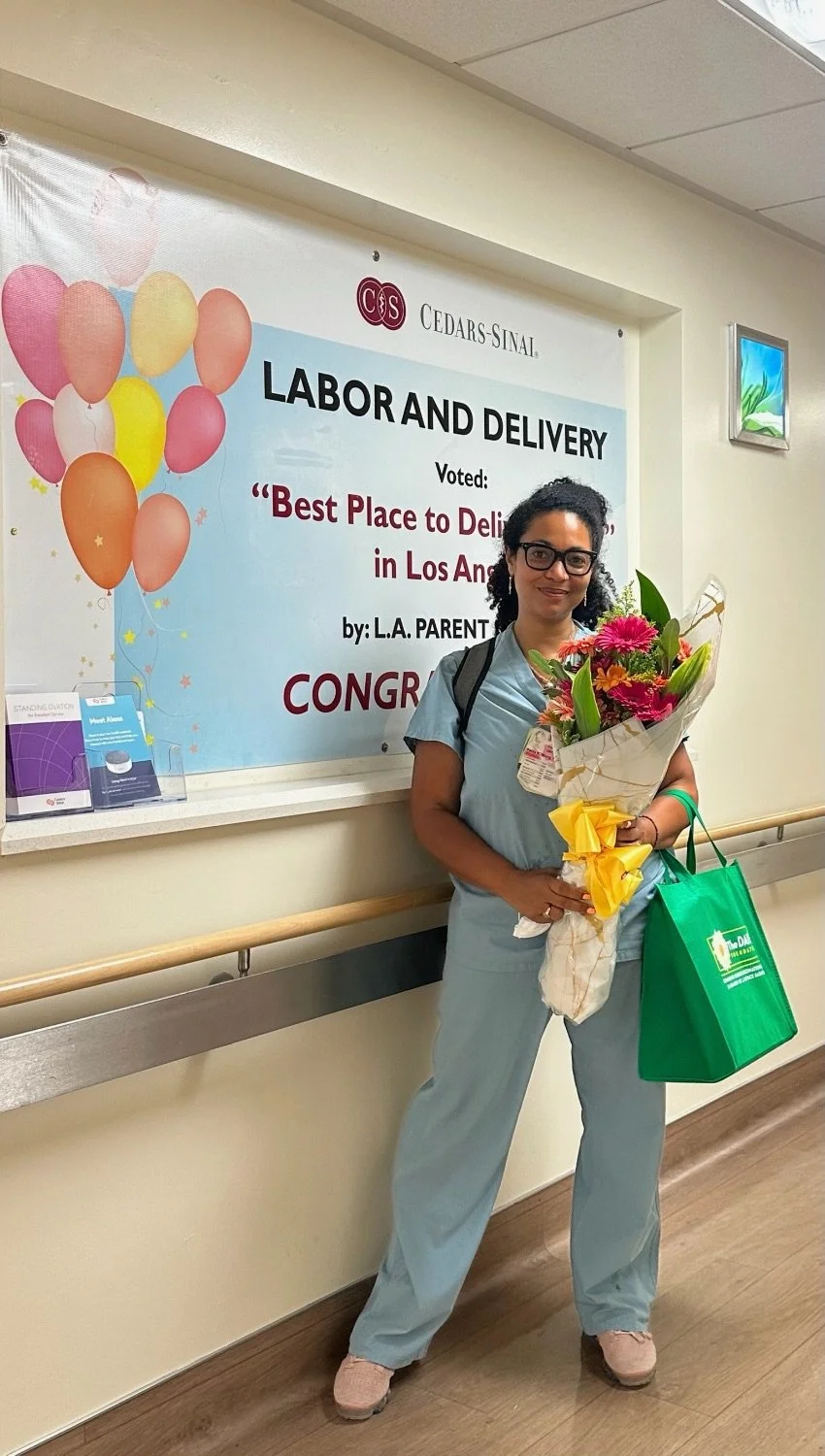 A woman in scrubs holding a bouquet of flowers and a green gift bag standing in front of a sign that recognizes her as the best place to deliver in Los Angeles, with colorful balloons on the sign.