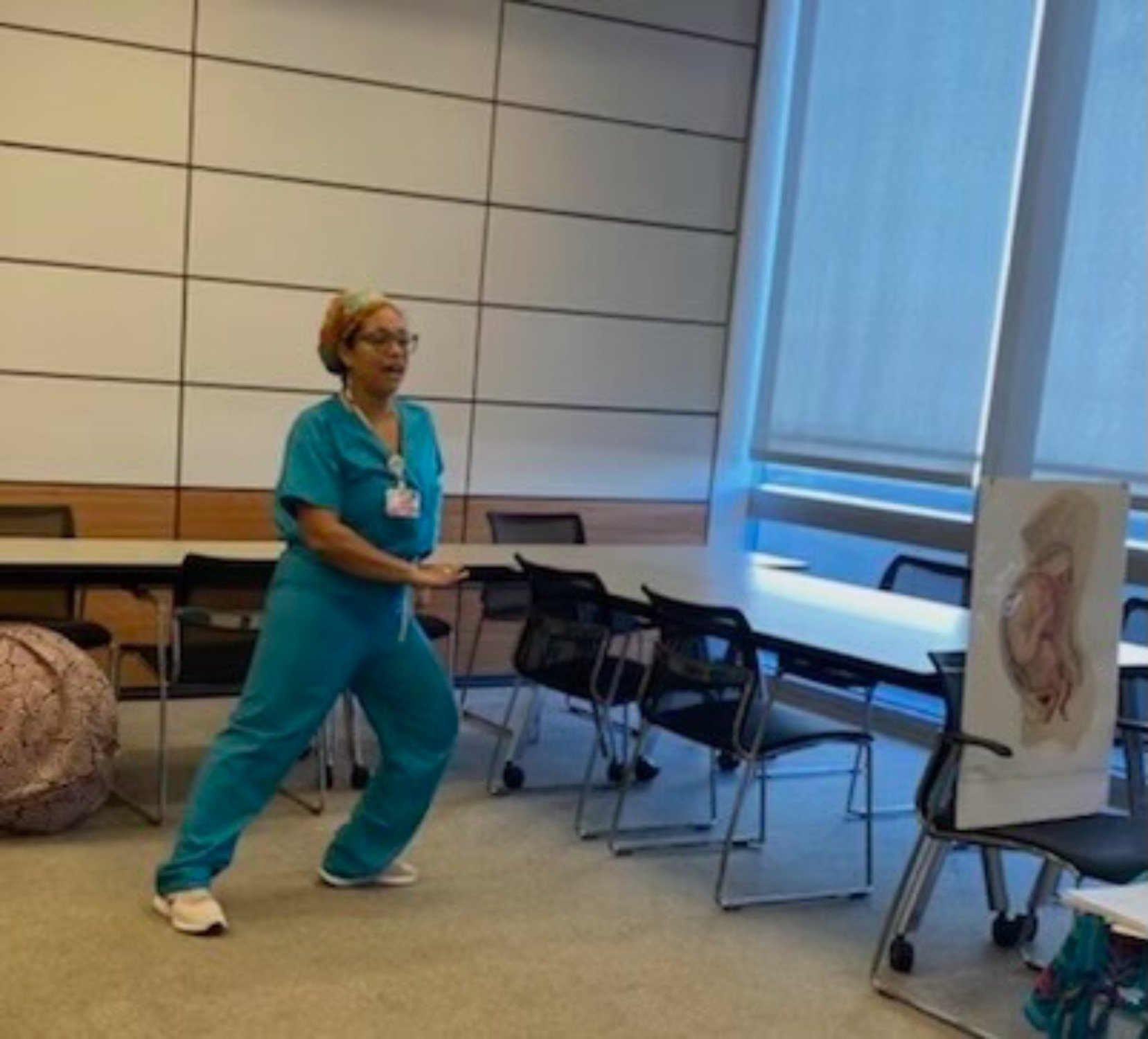 A woman dressed in teal scrubs performing a dance move in a room with chairs, a large window with blinds, and a medical illustration on an easel.