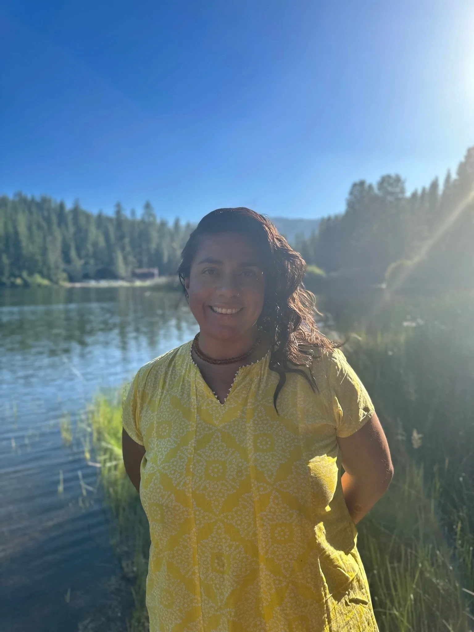 A woman in a yellow dress standing by a lake with trees and hills in the background on a sunny day.