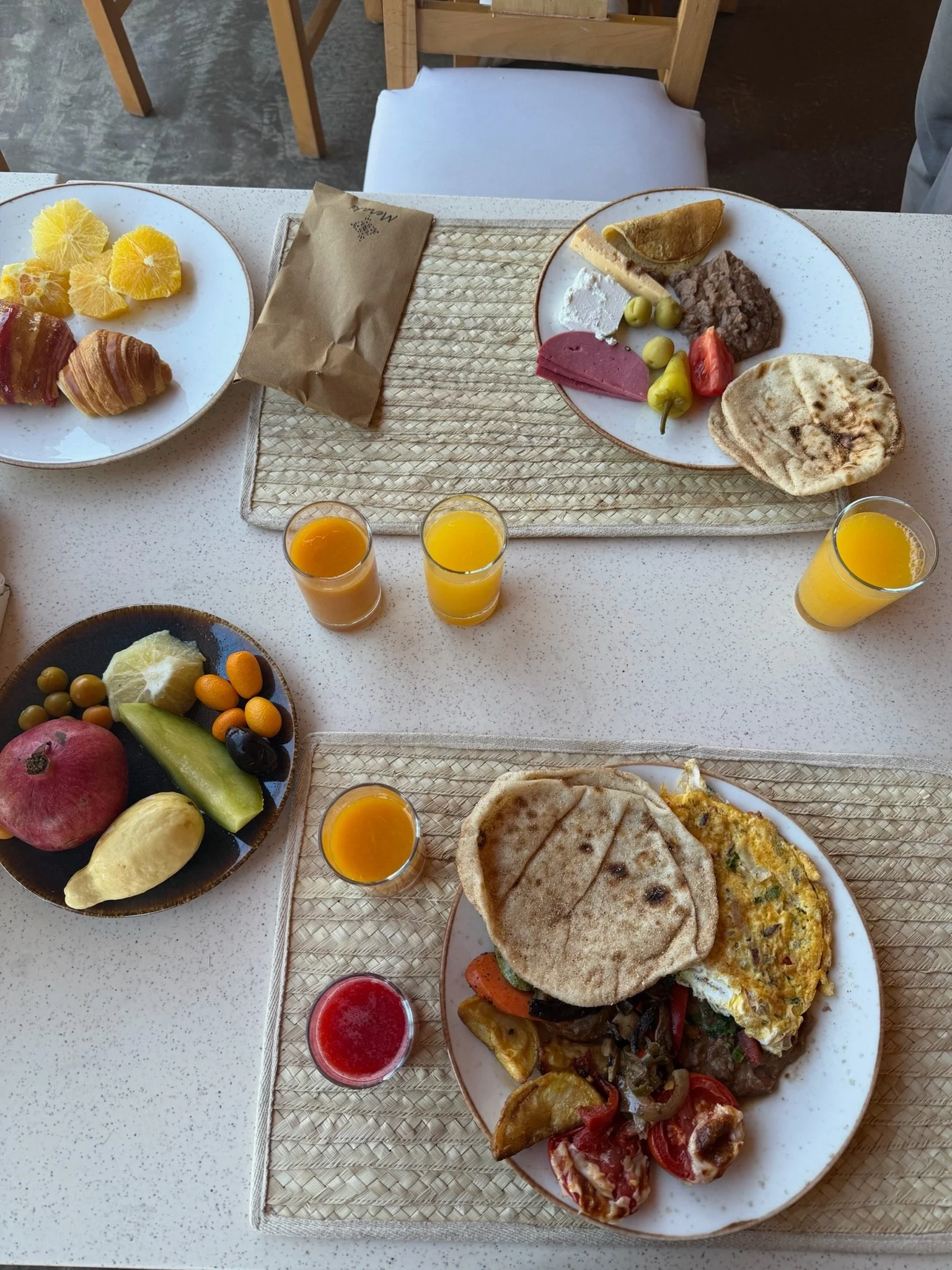 A breakfast spread featuring plates of fruit, pastries, a breakfast plate with eggs and vegetables, a plate with meats, cheese, olives, and bread, and glasses of orange juice and red drink on a white table.