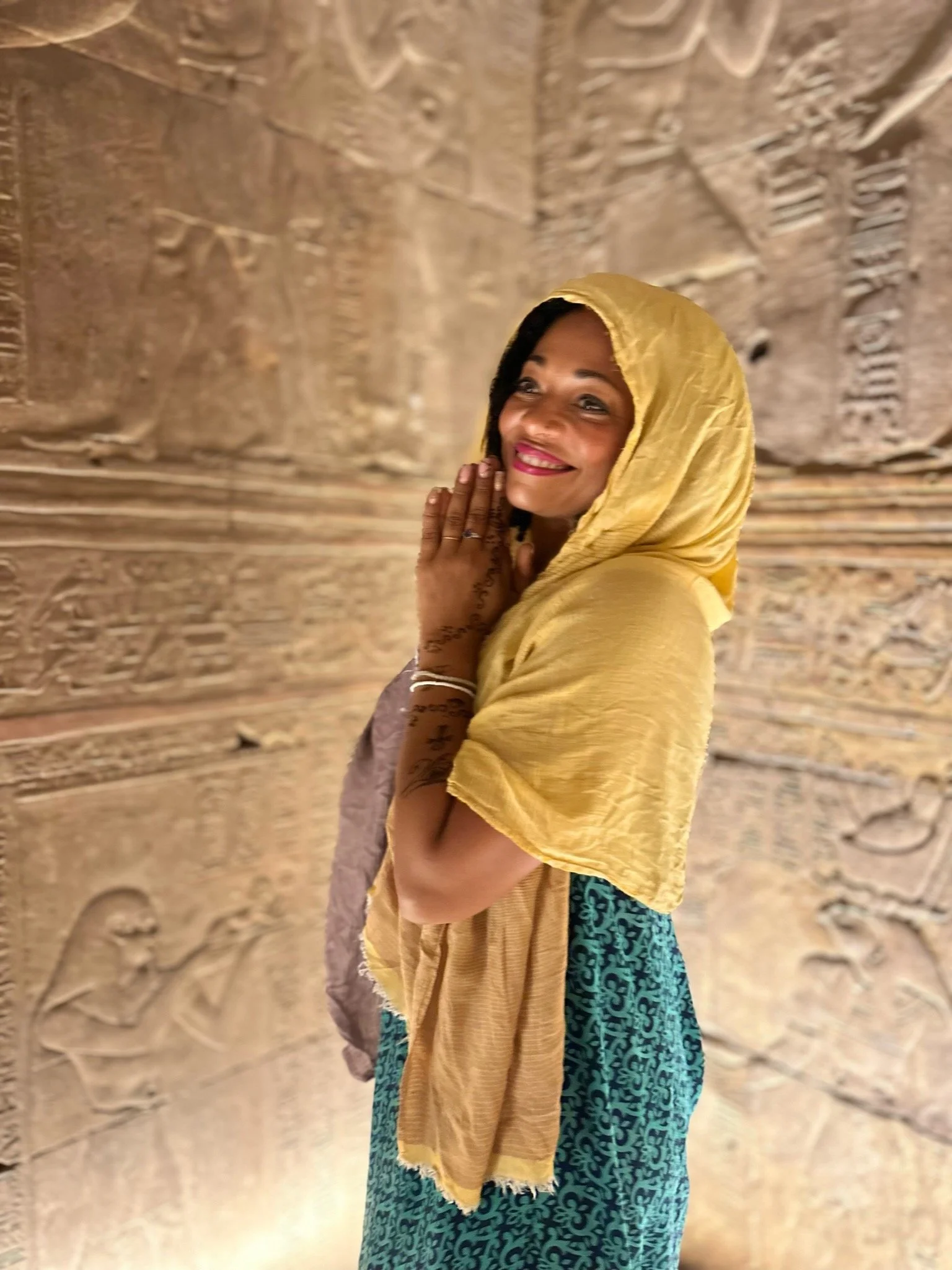 A woman with dark hair wearing a yellow headscarf, smiling, and clasping her hands in prayer position, standing in front of ancient carved stone hieroglyphics.
