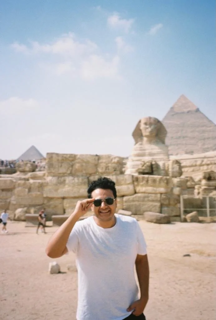 Ahmed, travel agent, the wearing sunglasses and a white t-shirt standing in front of the Great Sphinx of Giza and pyramids in Egypt on a sunny day.