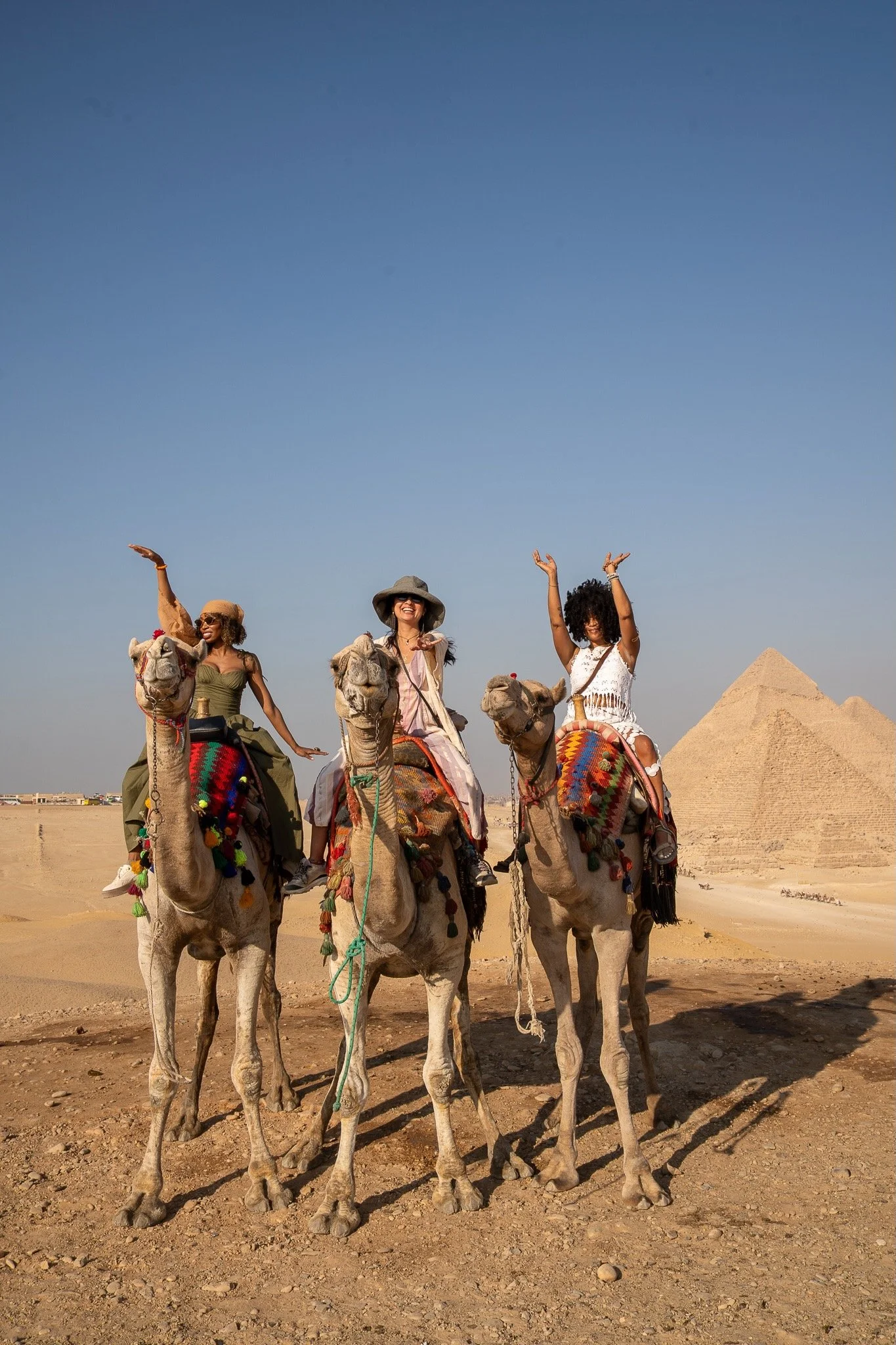 Three women from one of the retreats riding camels in a desert with a pyramid in the background.